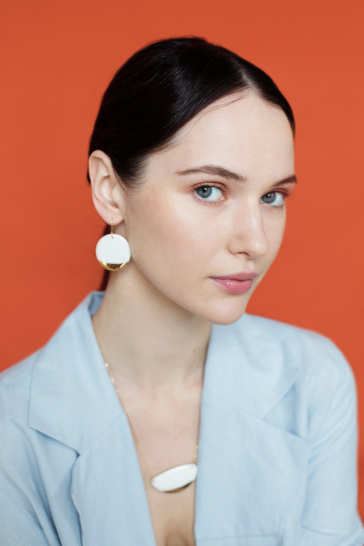 A woman with fair skin and blue eyes poses against an orange background, wearing a light blue blazer, her dark hair pulled back, and accessorized with Rozenthal Jewelrys Round Earrings.