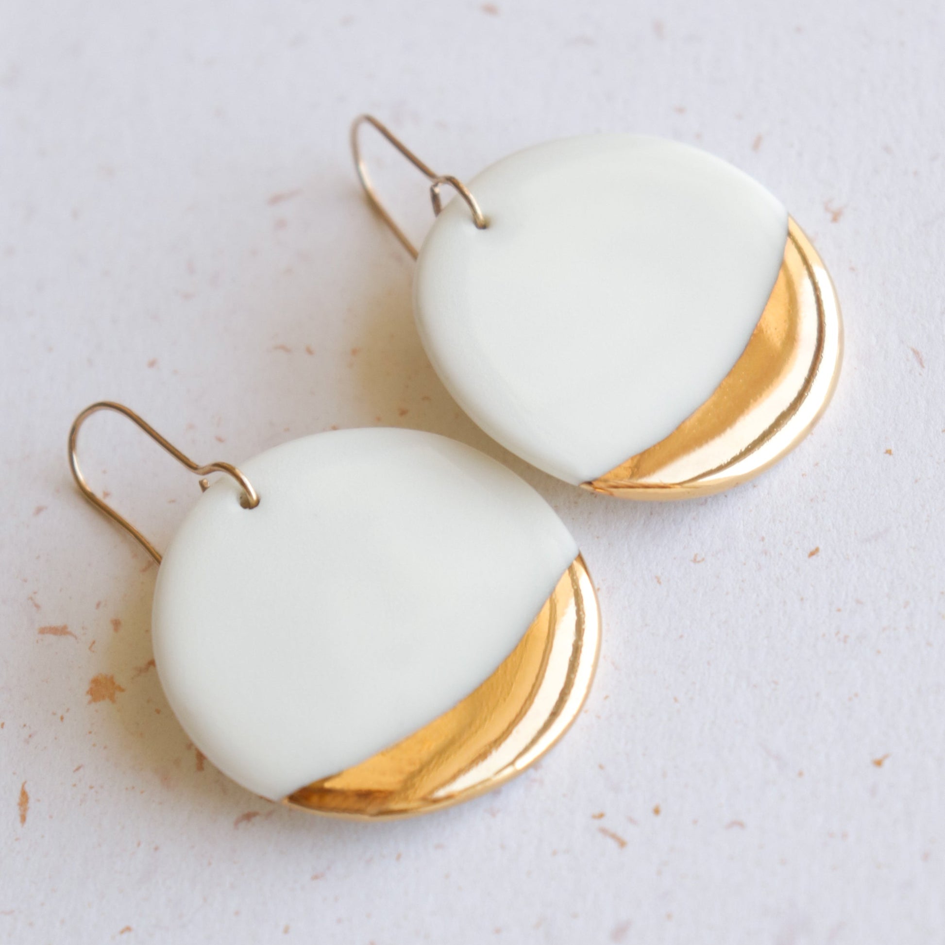 Rozenthal Jewelry’s Round Earrings feature a white porcelain upper half and shiny gold lower half, hanging from simple metal hooks. Shown on a light speckled surface, this minimalist design offers an elegant touch.