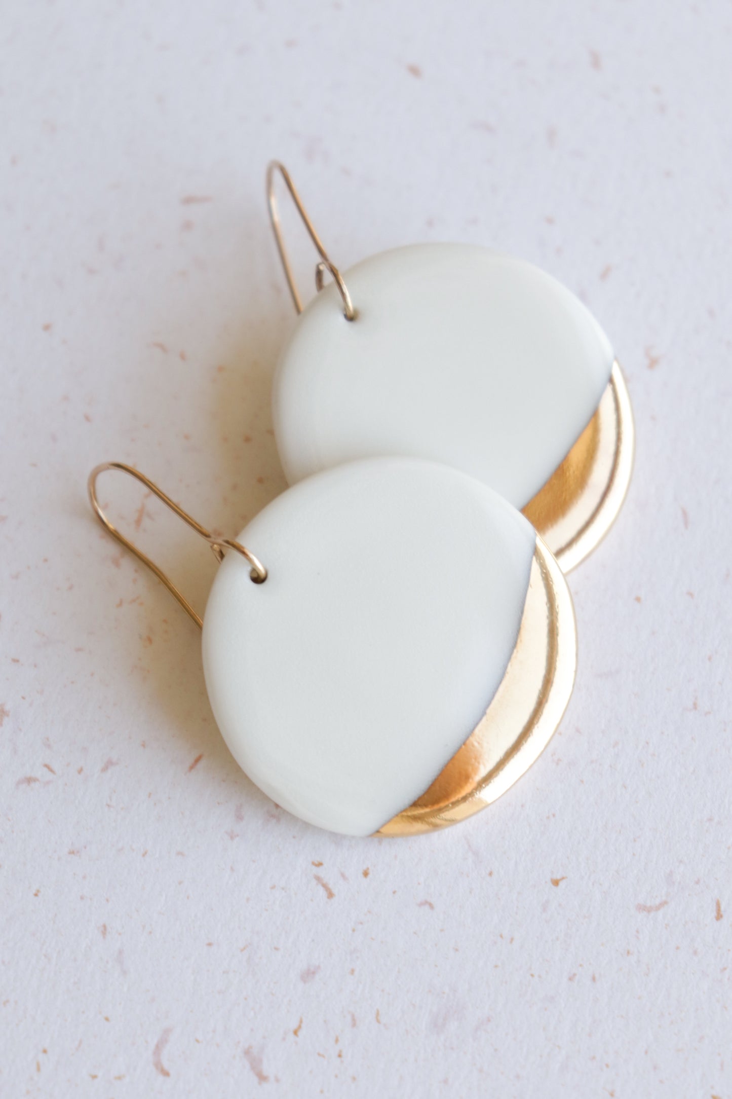The Round Earrings by Rozenthal Jewelry are white porcelain with gold accents and simple gold hooks, displayed on a light speckled surface—ideal for fans of minimalist jewelry.