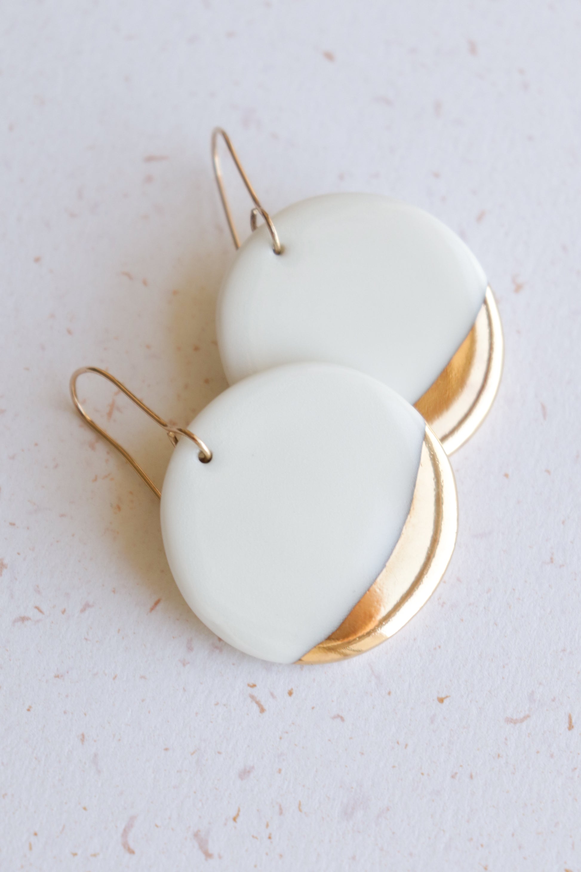 The Round Earrings by Rozenthal Jewelry are white porcelain with gold accents and simple gold hooks, displayed on a light speckled surface—ideal for fans of minimalist jewelry.