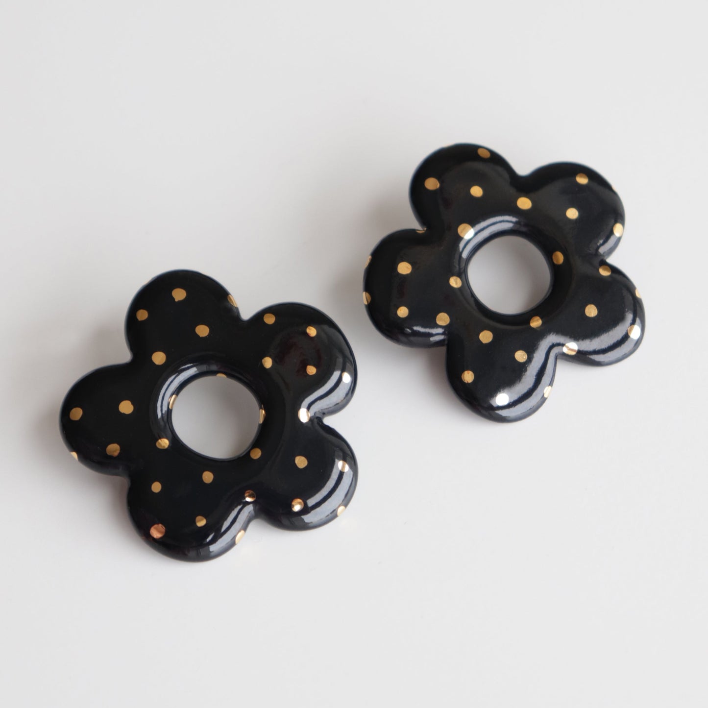 The Flower Earrings by Rozenthal Jewelry are glossy black porcelain, flower-shaped statement pieces with gold polka dots, each handmade with a circular center hole and displayed on a white surface.
