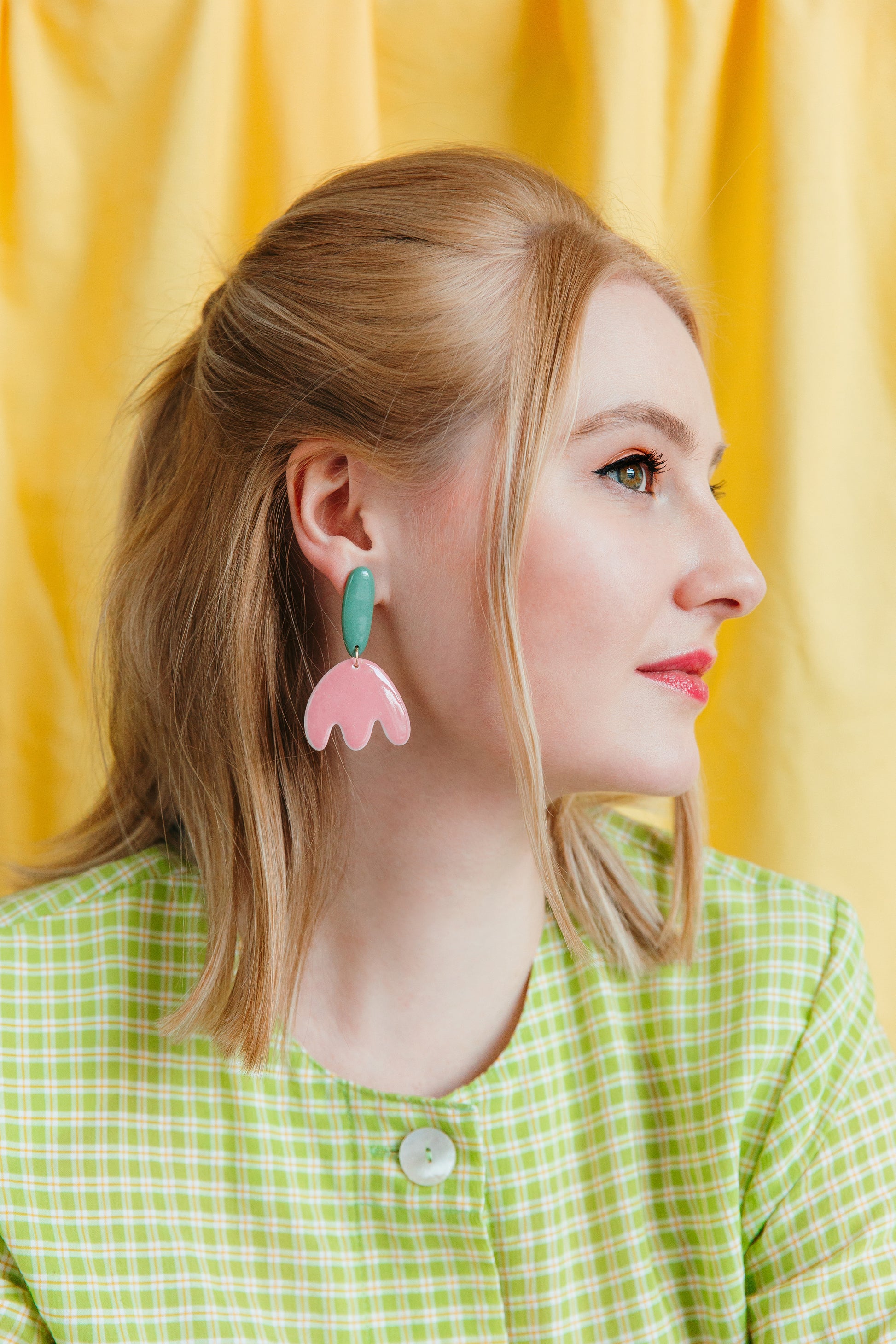 A blonde woman wearing a green plaid jacket and Rozenthal Jewelry’s Tulip Earrings gazes right, set against a yellow curtain backdrop.
