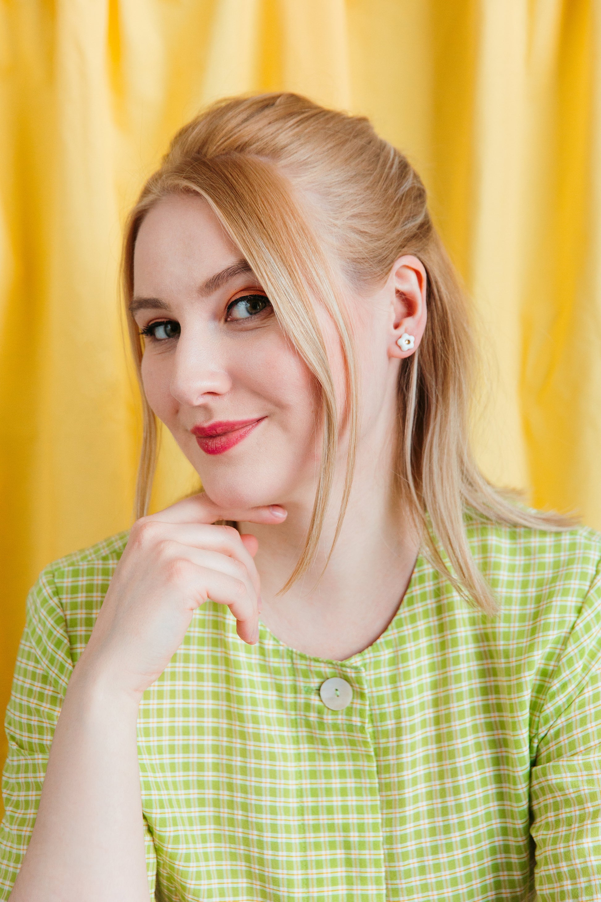 A woman with light skin and blonde hair wears Rozenthal Jewelrys Daisy Studs (XS) and a green and white checkered top, smiling gently with her hand on her chin against a bright yellow background.