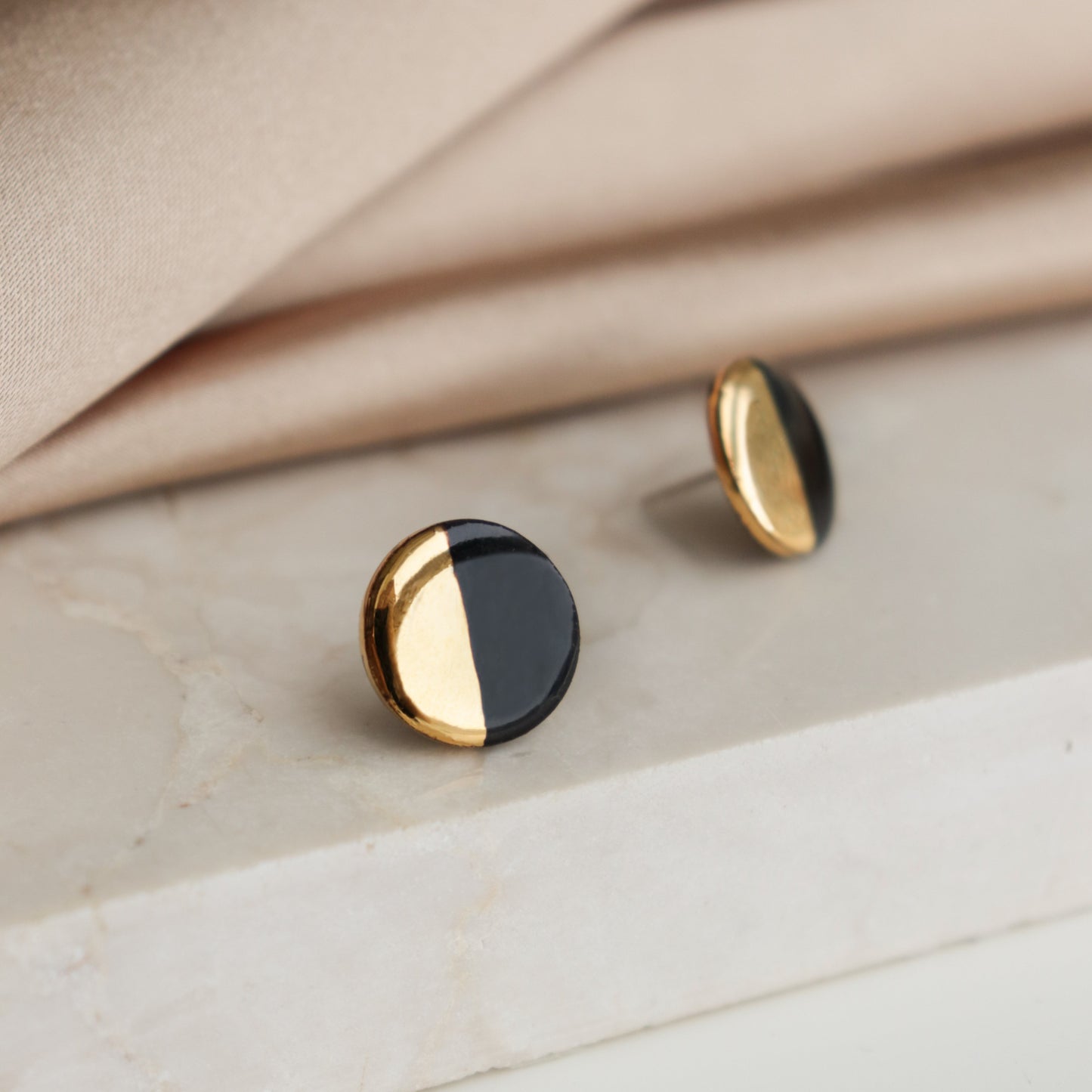The Rozenthal Jewelry Round Studs / S feature a black and gold half-and-half design, shown on white marble with beige fabric—ideal for fans of minimalist jewelry.