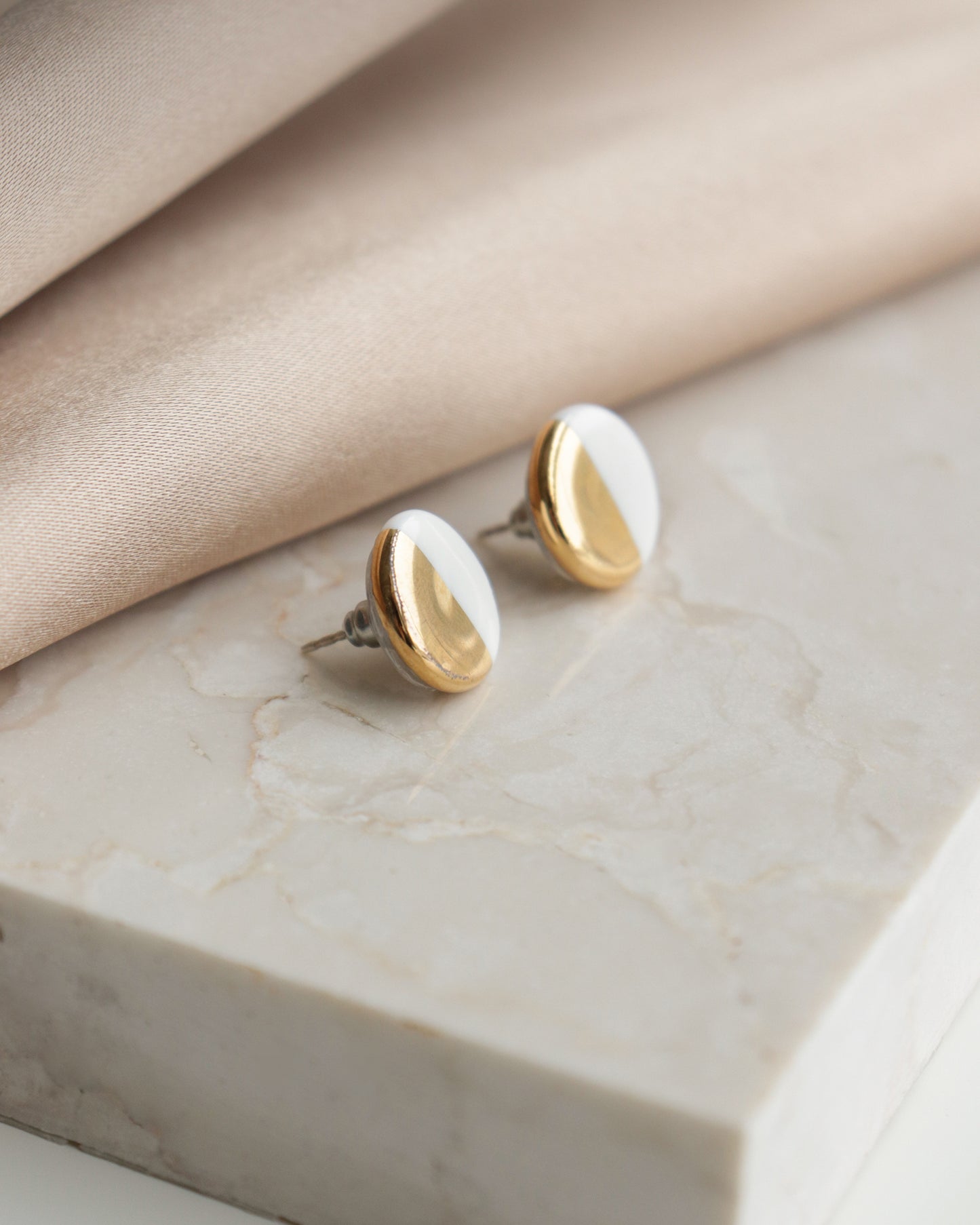 Round Studs / S by Rozenthal Jewelry feature a gold and white half-and-half design, shown on a white marble surface beside beige fabric—ideal for those who appreciate minimalist jewelry.