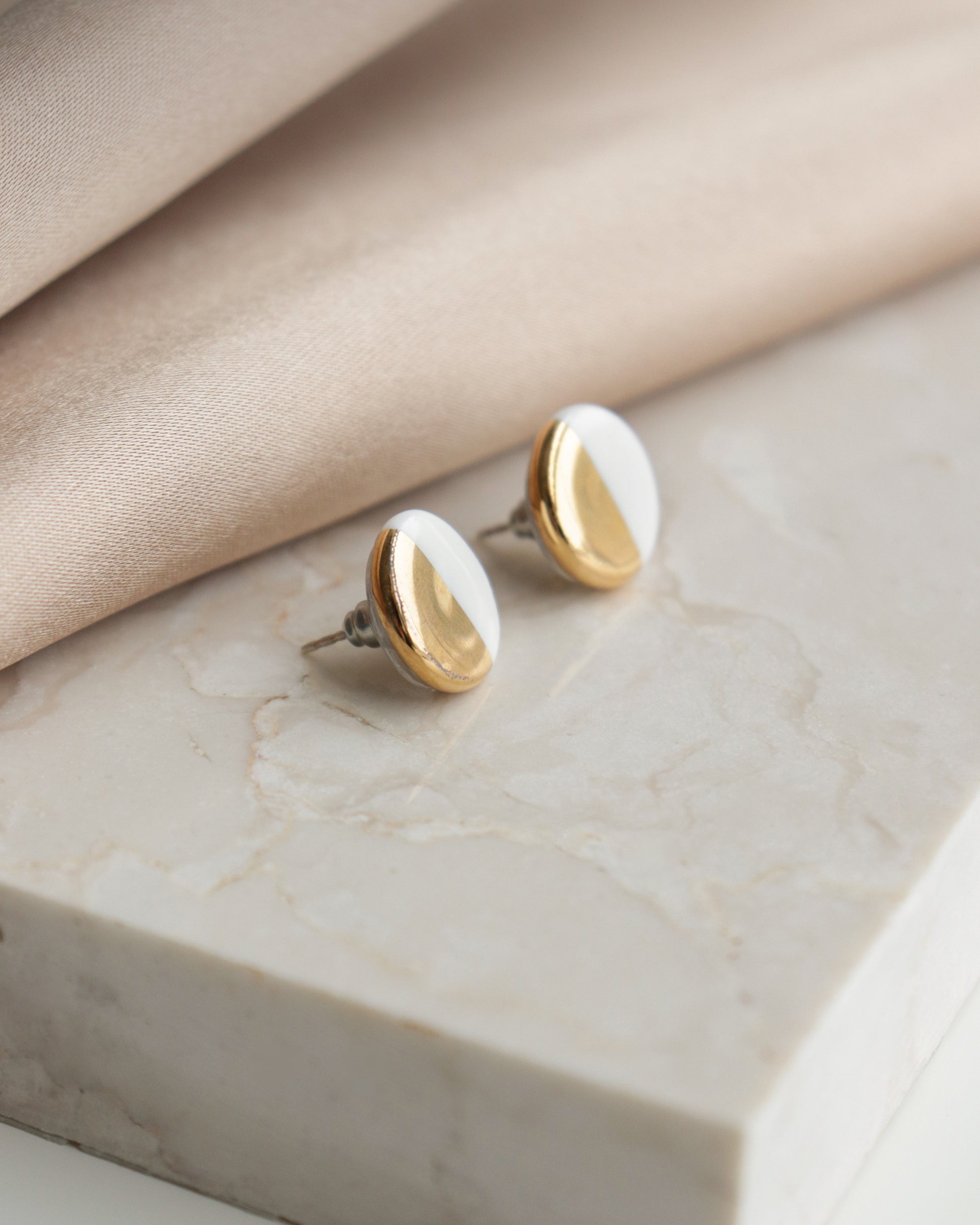 Round Studs / S by Rozenthal Jewelry feature a gold and white half-and-half design, shown on a white marble surface beside beige fabric—ideal for those who appreciate minimalist jewelry.