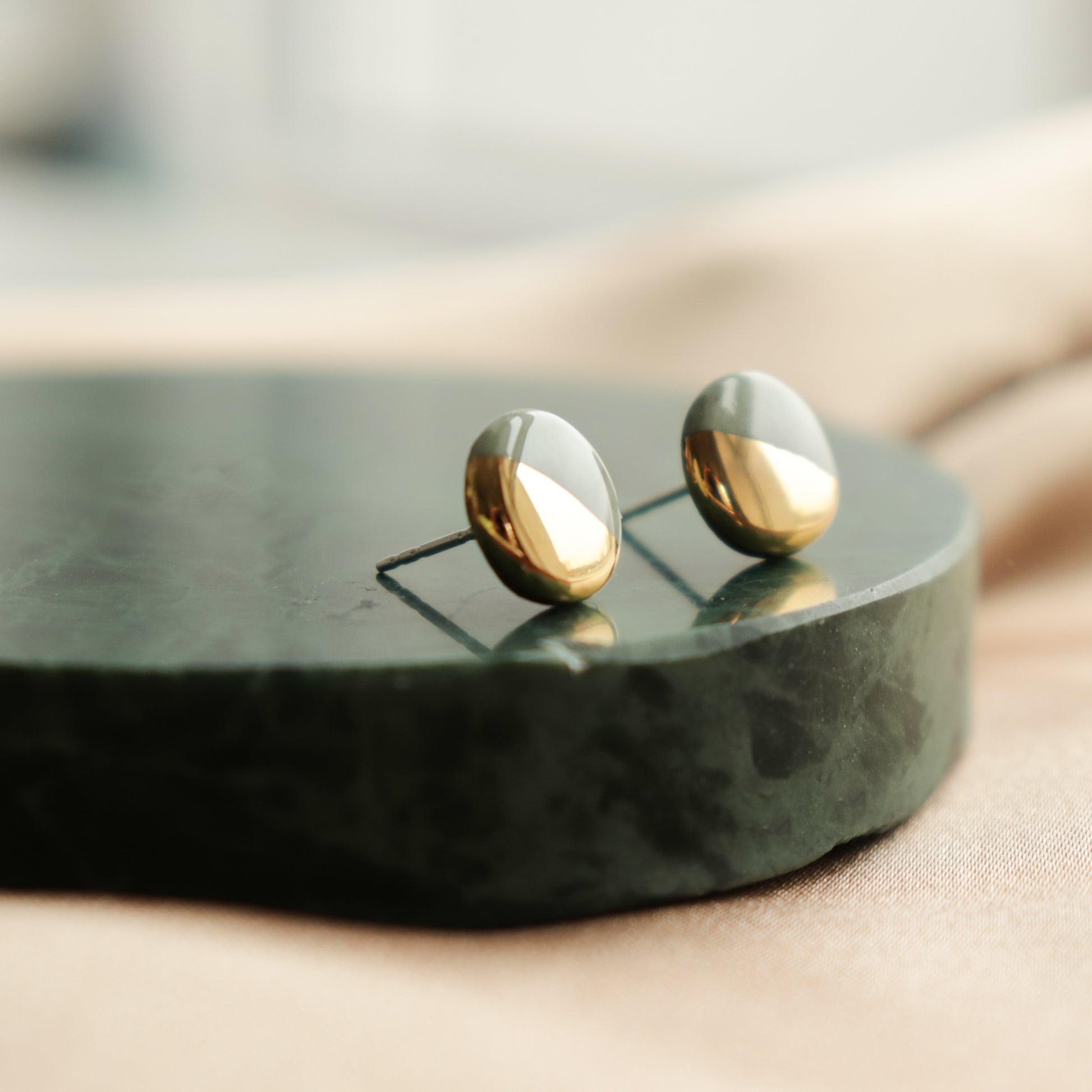Rozenthal Jewelrys Round Studs / XS in gold and silver are ideal for minimalist jewelry lovers, shown on a dark green circular marble slab against a soft beige fabric background.