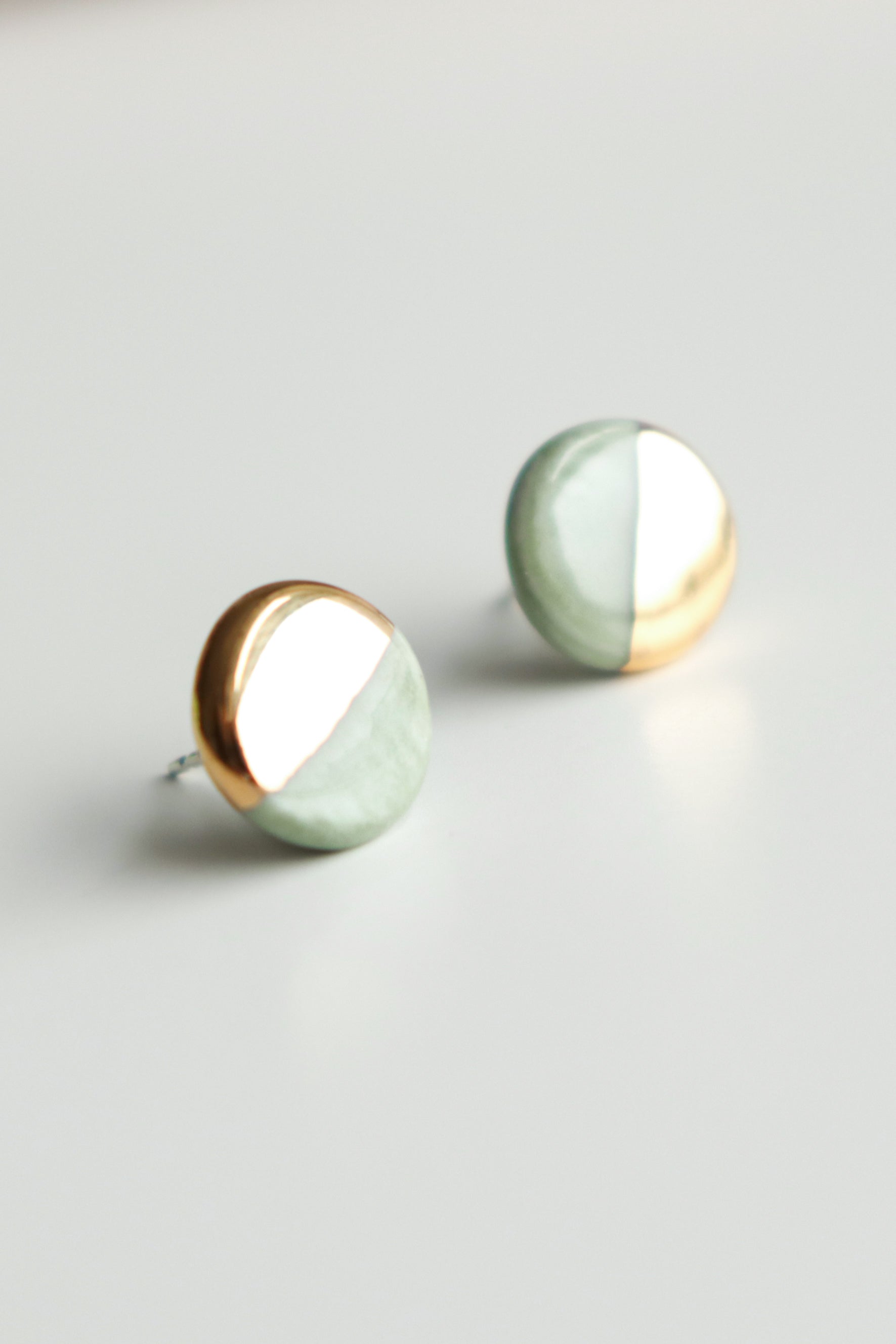 Handmade by Rozenthal Jewelry, the Round Studs / XS are minimalist porcelain earrings with a half gold metallic and half light green matte finish, elegantly displayed on a white surface.