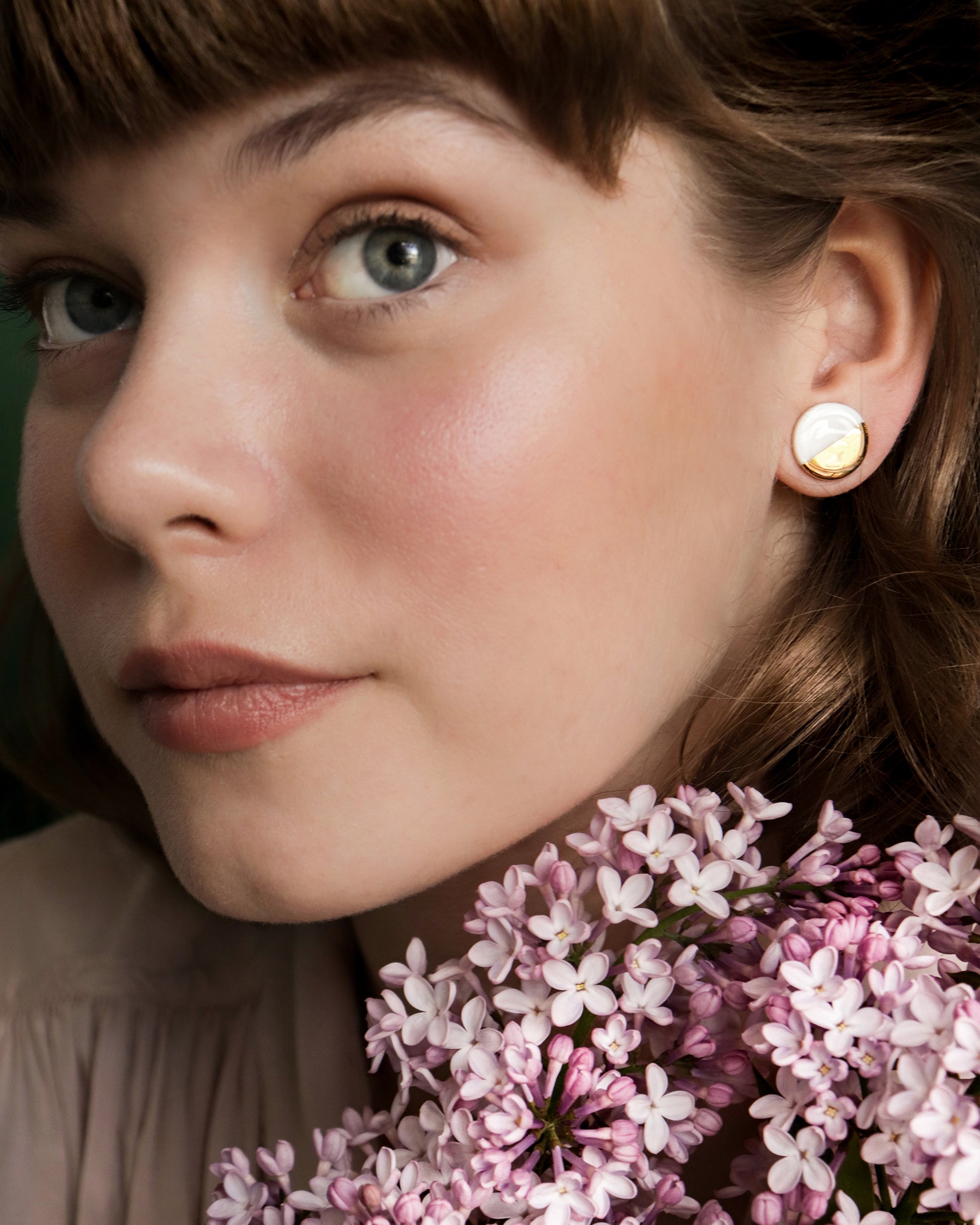 A woman with light skin and blue eyes holds lilac flowers near her face. Her brown hair has bangs, and she wears Rozenthal Jewelrys Round Studs / S—a minimalist gold earring with a white stone. Her makeup is natural and soft.