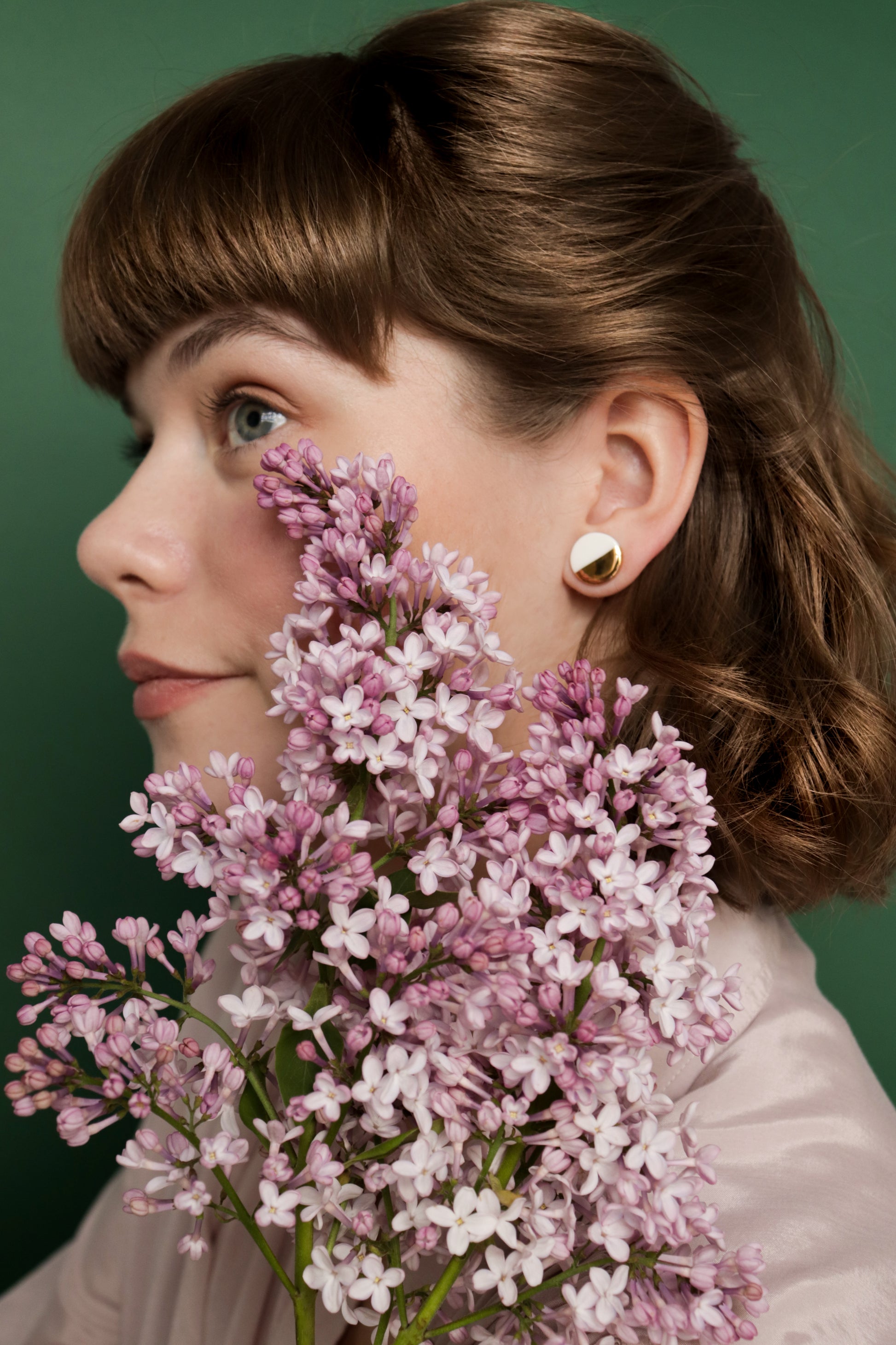 A woman with light skin and brown hair wears Rozenthal Jewelry Round Studs / S minimalist gold earrings, posing in front of a green background while holding a large bouquet of light purple lilac flowers near her face.