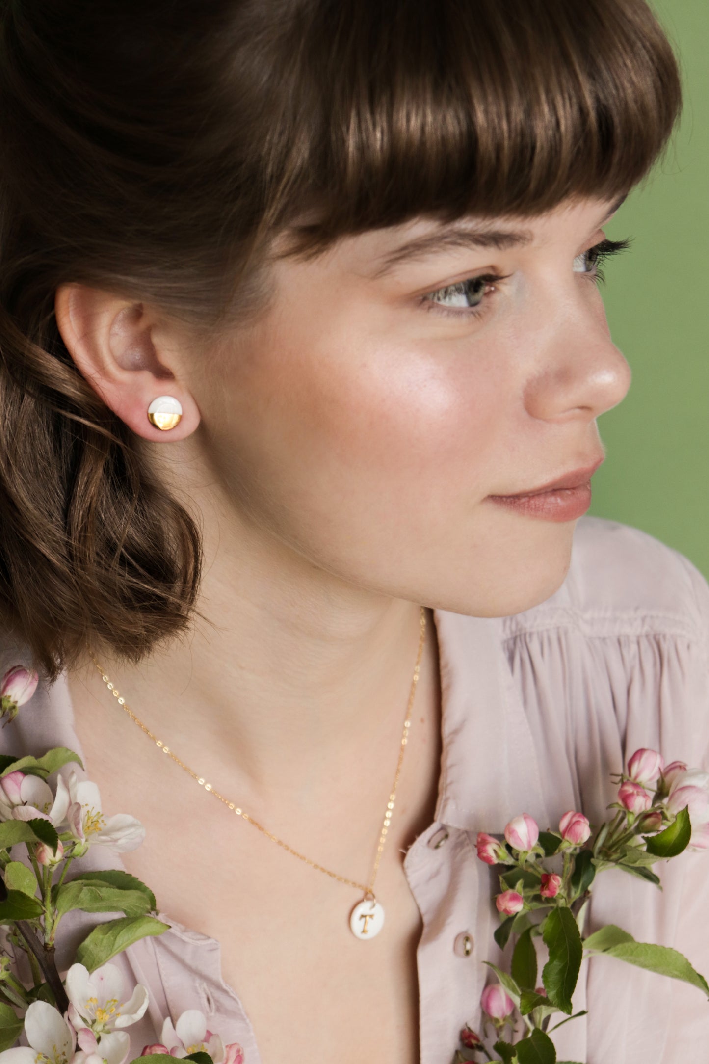 A woman with light brown hair and bangs wears minimalist jewelry—Rozenthal Jewelrys Round Studs / XS and a gold necklace. She holds pink and white blossoms, gazing to the side against a green background.