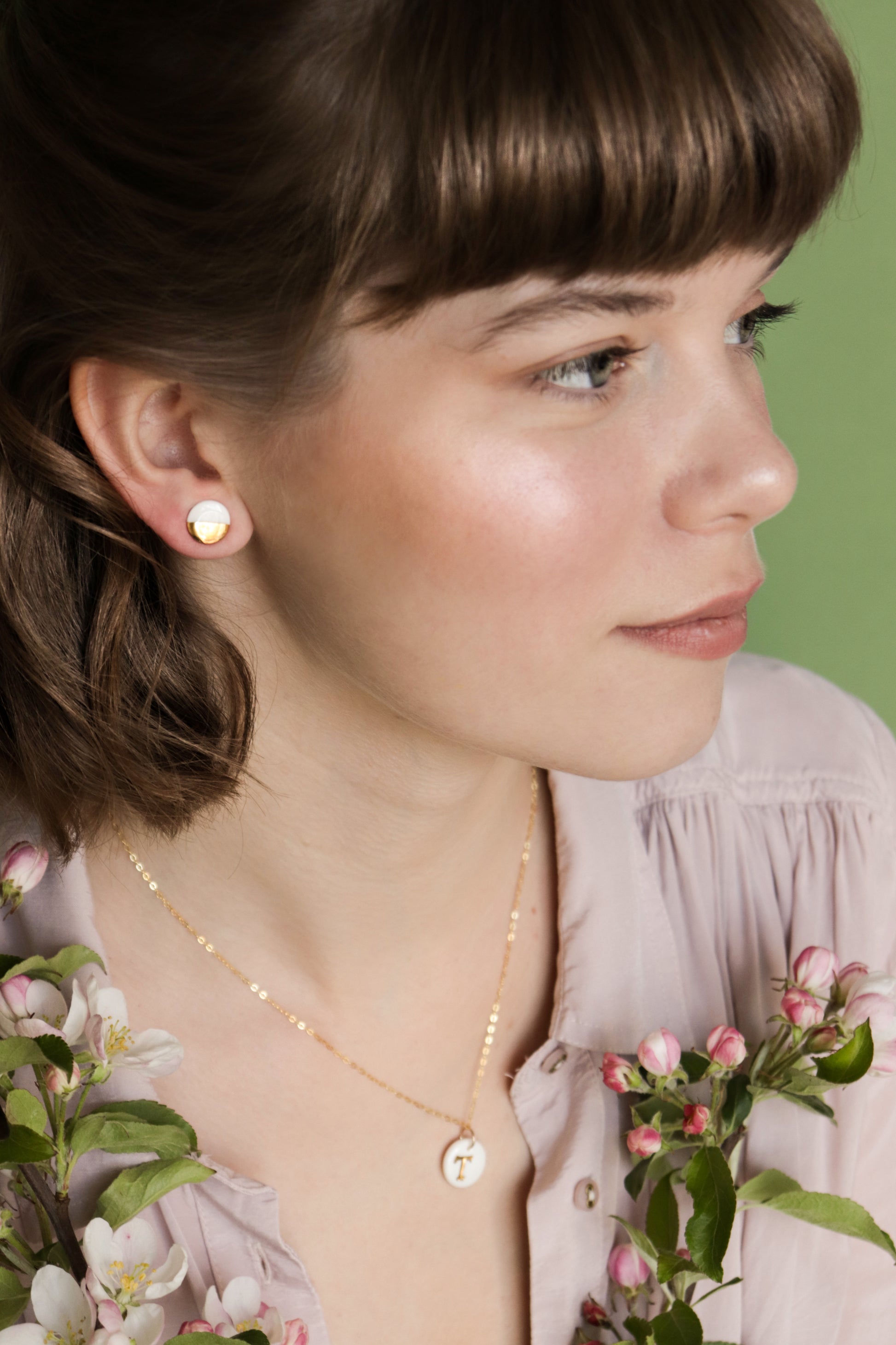 A woman with light brown hair and bangs wears minimalist jewelry—Rozenthal Jewelrys Round Studs / XS and a gold necklace. She holds pink and white blossoms, gazing to the side against a green background.