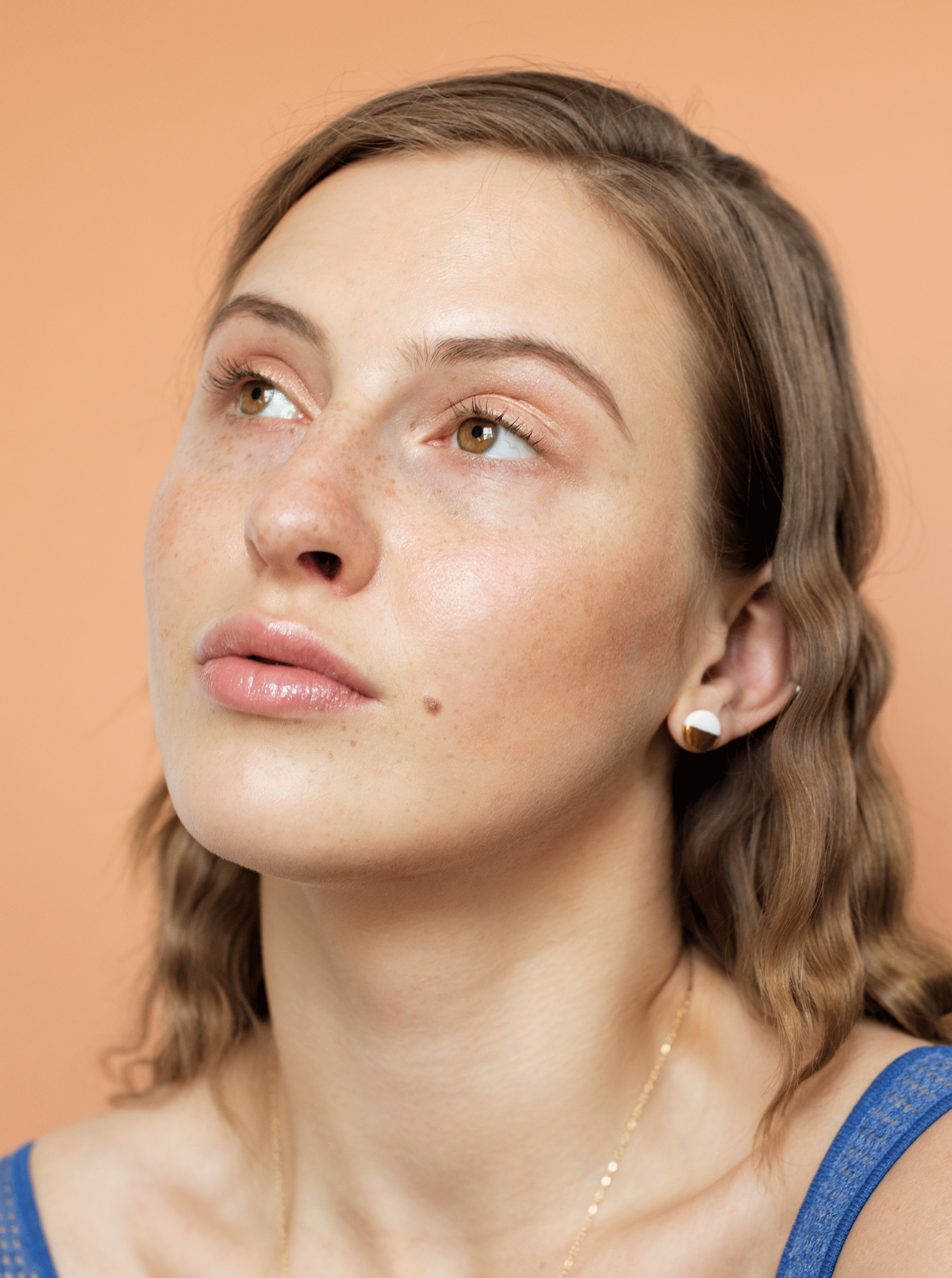 A young woman with wavy light brown hair, clear skin, freckles, and minimal makeup wears a blue top and Rozenthal Jewelrys Round Studs / XS gold earrings, plus a delicate porcelain necklace, against a peach-colored background.