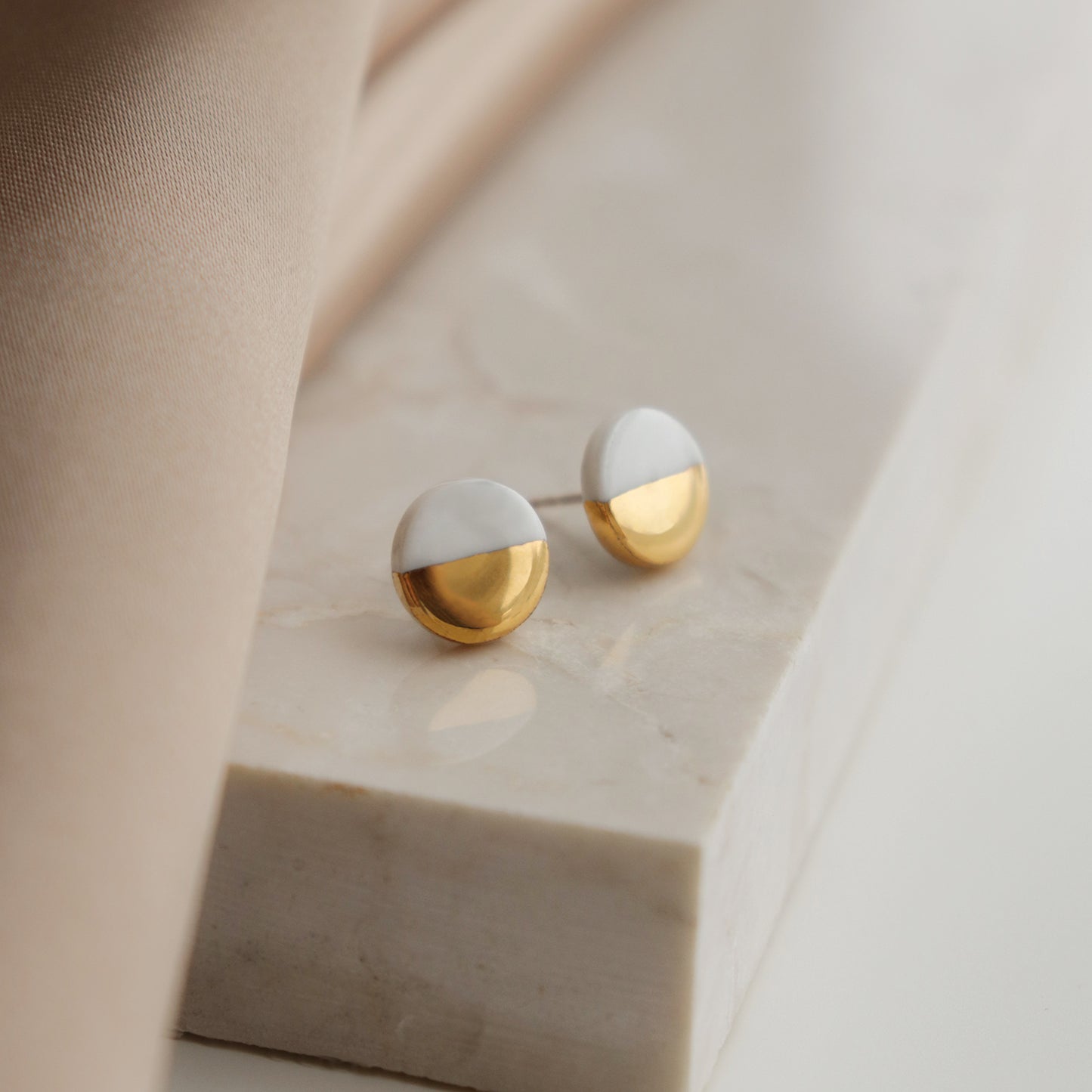 A pair of Round Studs / XS by Rozenthal Jewelry, featuring a minimalist half-white, half-gold design, rests on a marble surface next to beige fabric.