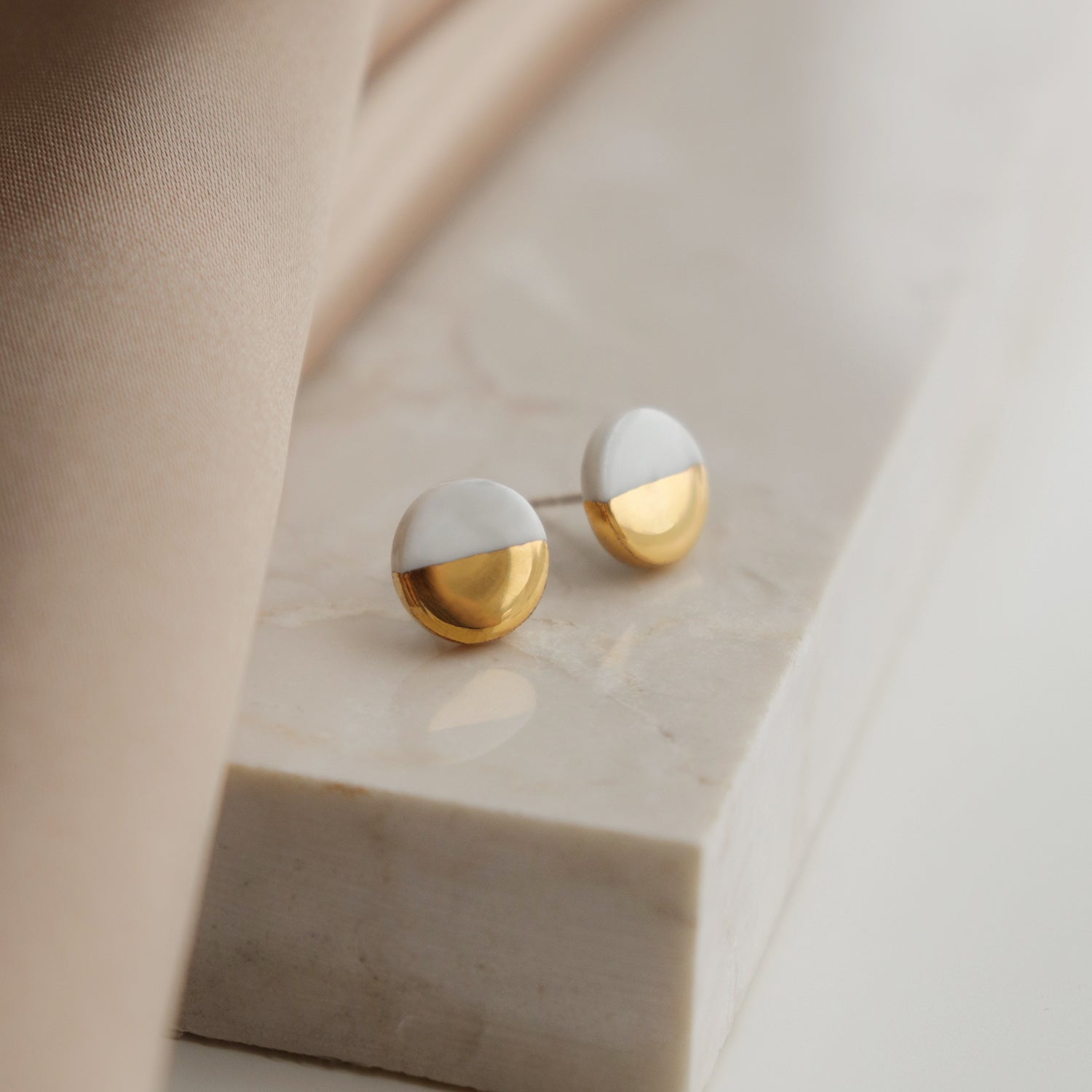 A pair of Round Studs / XS by Rozenthal Jewelry, featuring a minimalist half-white, half-gold design, rests on a marble surface next to beige fabric.