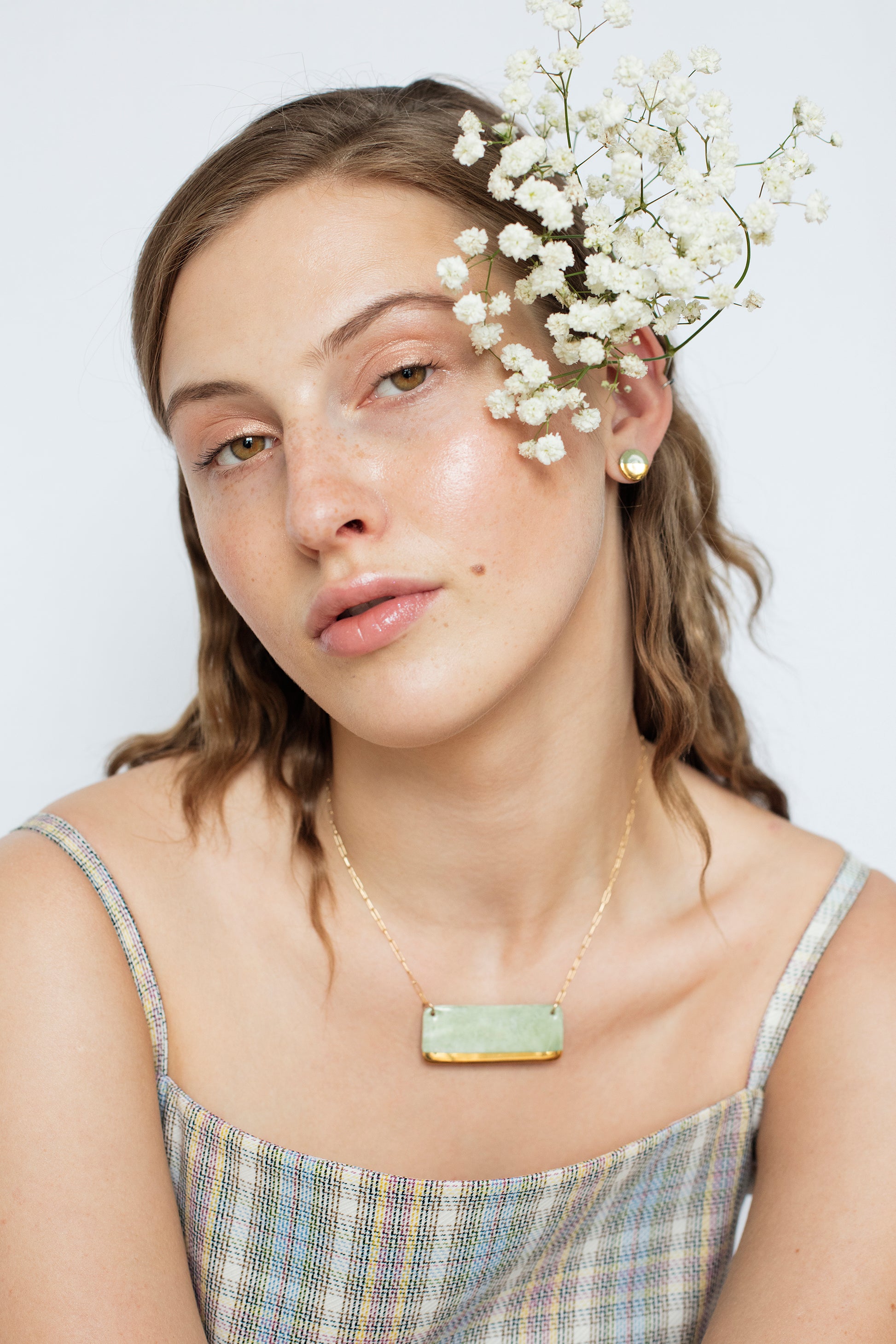 A young woman with light brown hair and freckles wears Rozenthal Jewelrys Round Studs / XS, a sleeveless plaid top, minimalist jewelry, and babys breath flowers behind her ear, posing against a plain white background.