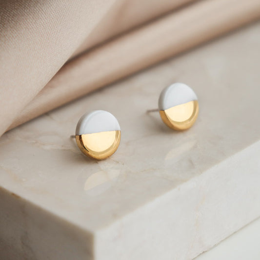 The Rozenthal Jewelry Round Studs / XS feature a minimalist design with white enamel on the top half and shiny gold on the bottom, shown on a light marble surface with beige fabric in the background.