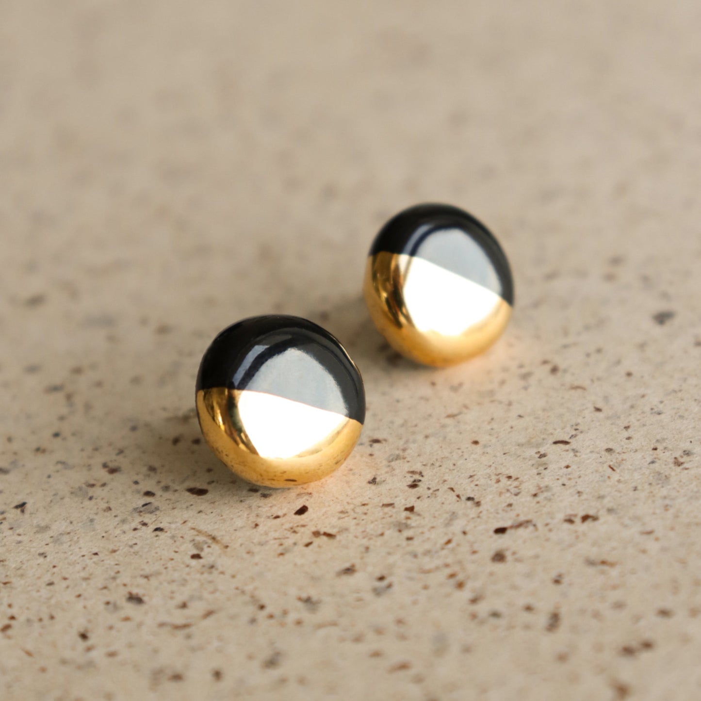 The Round Studs / XS by Rozenthal Jewelry feature a shiny gold lower half and glossy black upper half—perfect minimalist earrings, pictured on a speckled beige surface.