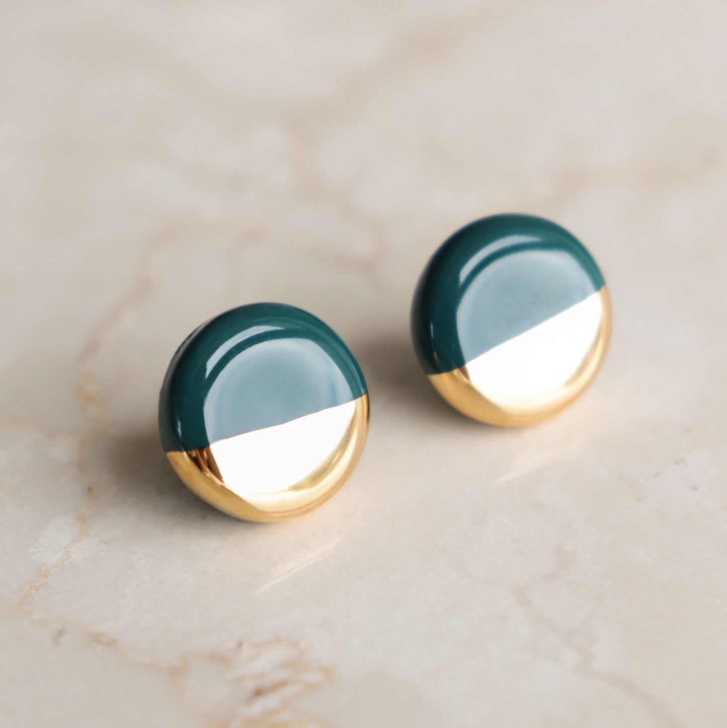 The Round Studs / S by Rozenthal Jewelry feature a minimalist half-dark green, half-gold design and are elegantly displayed on a light beige marble surface.