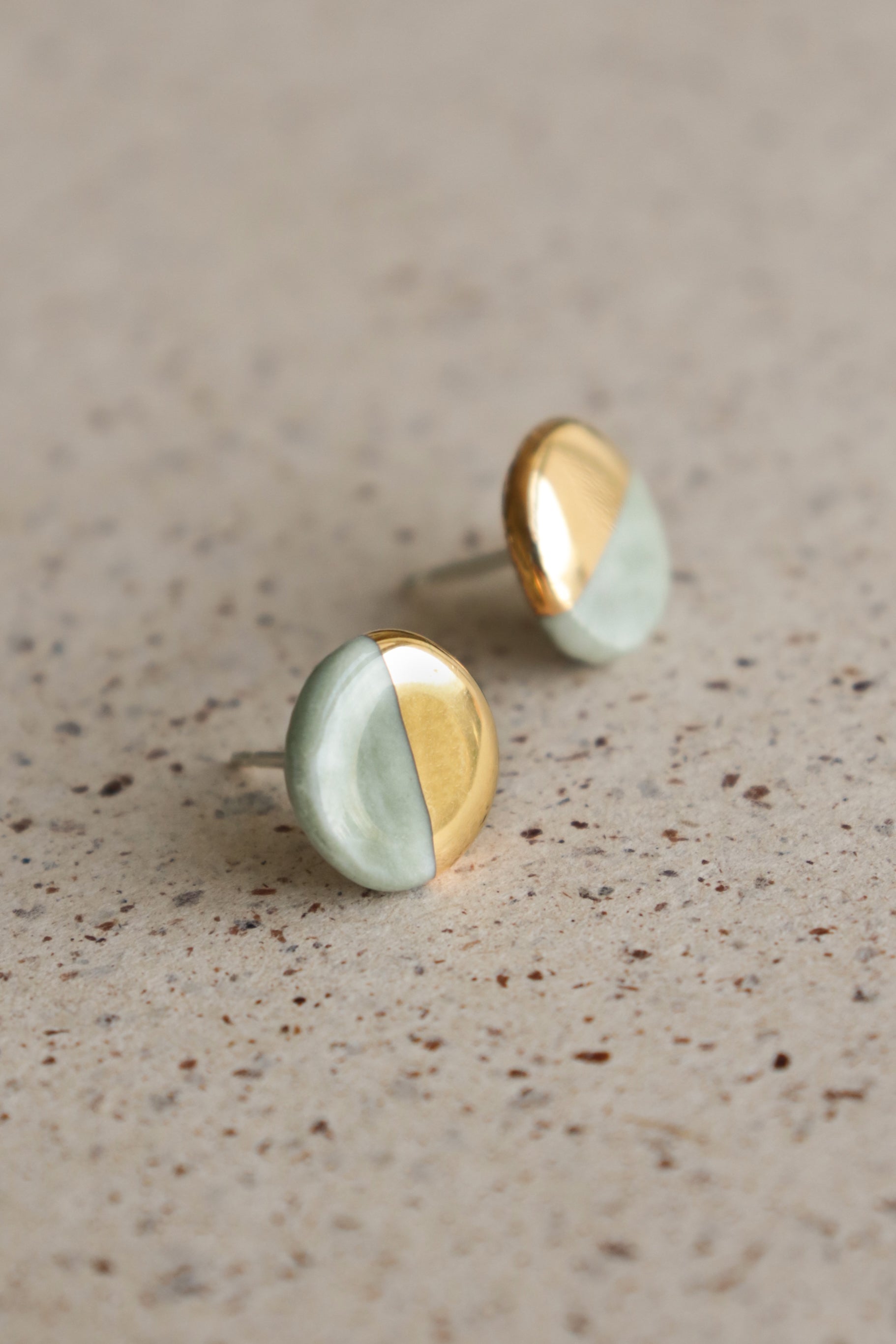 The Round Studs / XS by Rozenthal Jewelry feature a half-gold, half-mint green design and are displayed on a speckled beige surface—an elegant showcase of minimalist style.