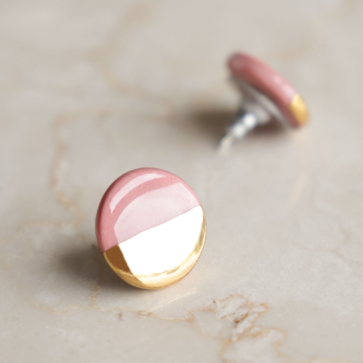 A pair of Rozenthal Jewelry Round Studs / S, featuring a pink upper half and shiny gold lower half, is shown on a light marble surface with one earring in sharp focus and the other softly blurred in the background.