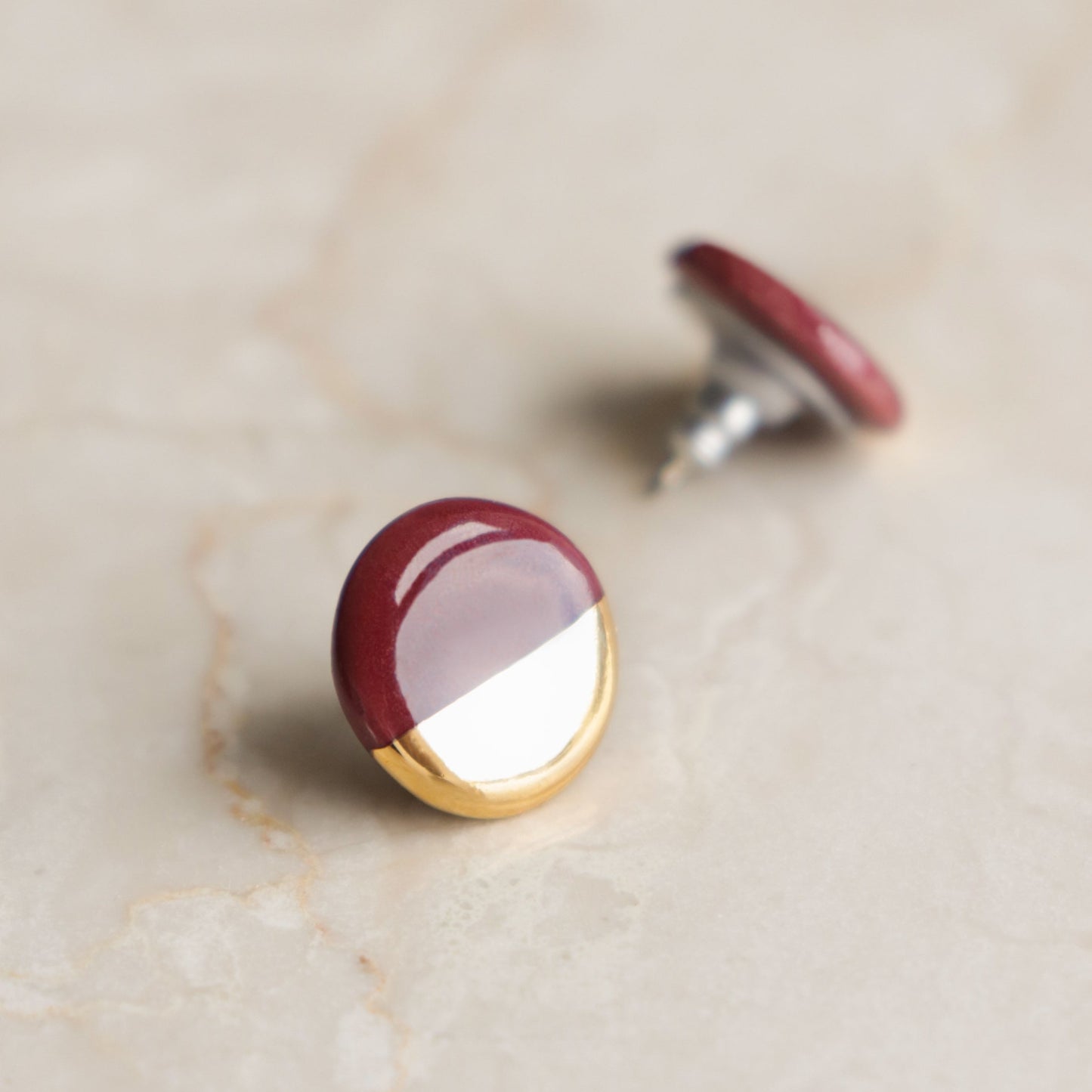 A close-up of Rozenthal Jewelrys Round Studs / S shows two minimalist earrings with glossy maroon tops, metallic gold bottoms, and silver posts, displayed on a light beige marble surface.