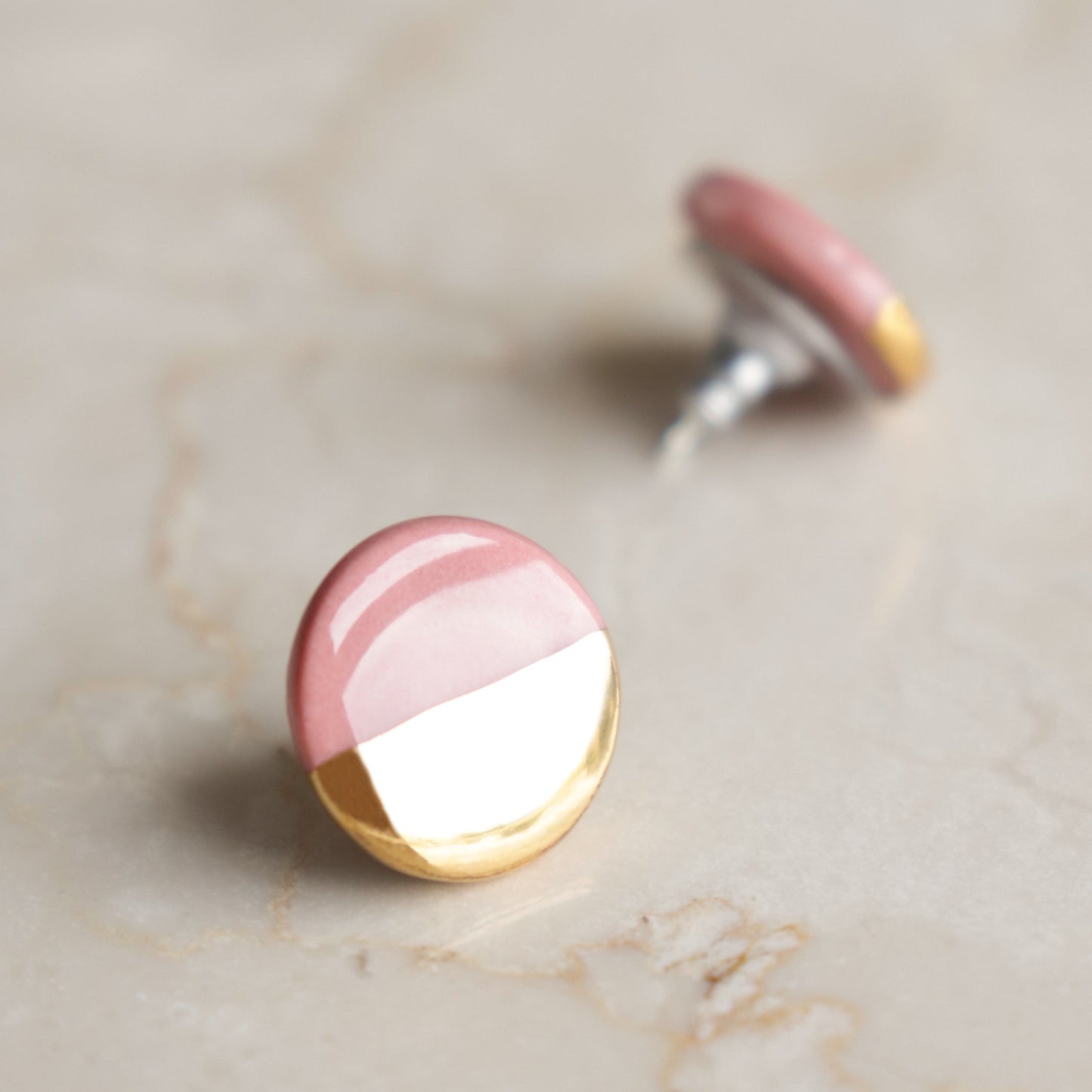 A close-up of Rozenthal Jewelry’s Round Studs / XS on marble, showing minimalist earrings with pink tops and gold bottoms. One porcelain earring is in focus while the other appears blurred in the background.
