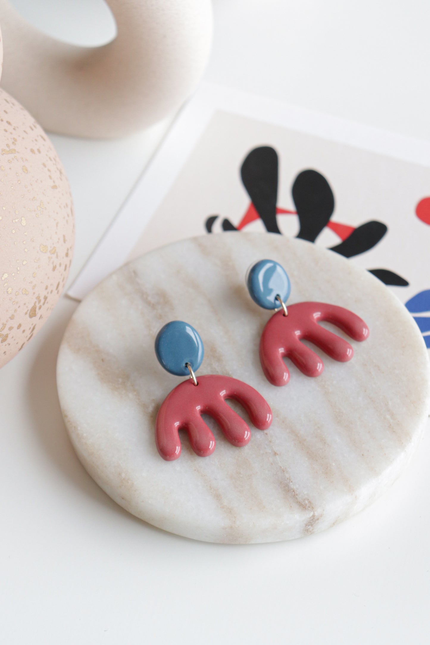 A pair of Rozenthal Jewelry Matisse Earrings, featuring lightweight waterproof blue circular tops and red abstract wavy shapes, rest on a round marble tray with modern decor and an abstract art print in the background.