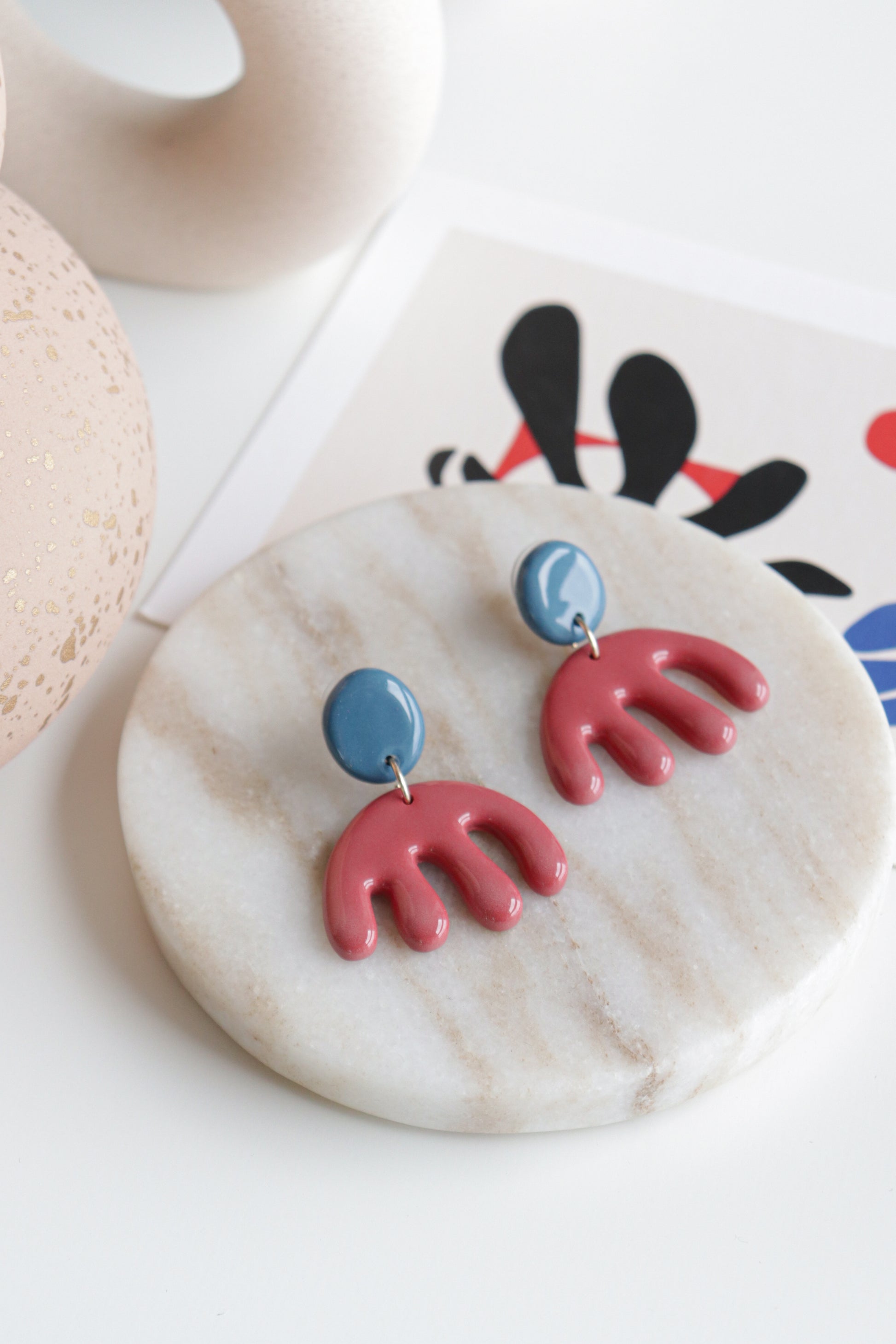 A pair of Rozenthal Jewelry Matisse Earrings, featuring lightweight waterproof blue circular tops and red abstract wavy shapes, rest on a round marble tray with modern decor and an abstract art print in the background.
