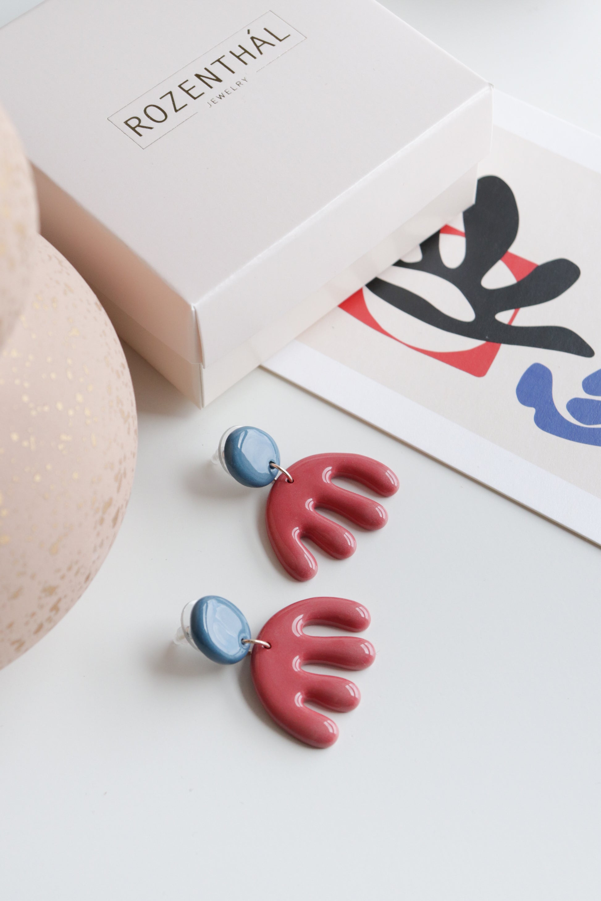 The Matisse Earrings by Rozenthal Jewelry, featuring blue circular tops and red abstract hand-shaped dangles, are displayed on a white surface near a decorative card and a box labeled ROZENTHAL.