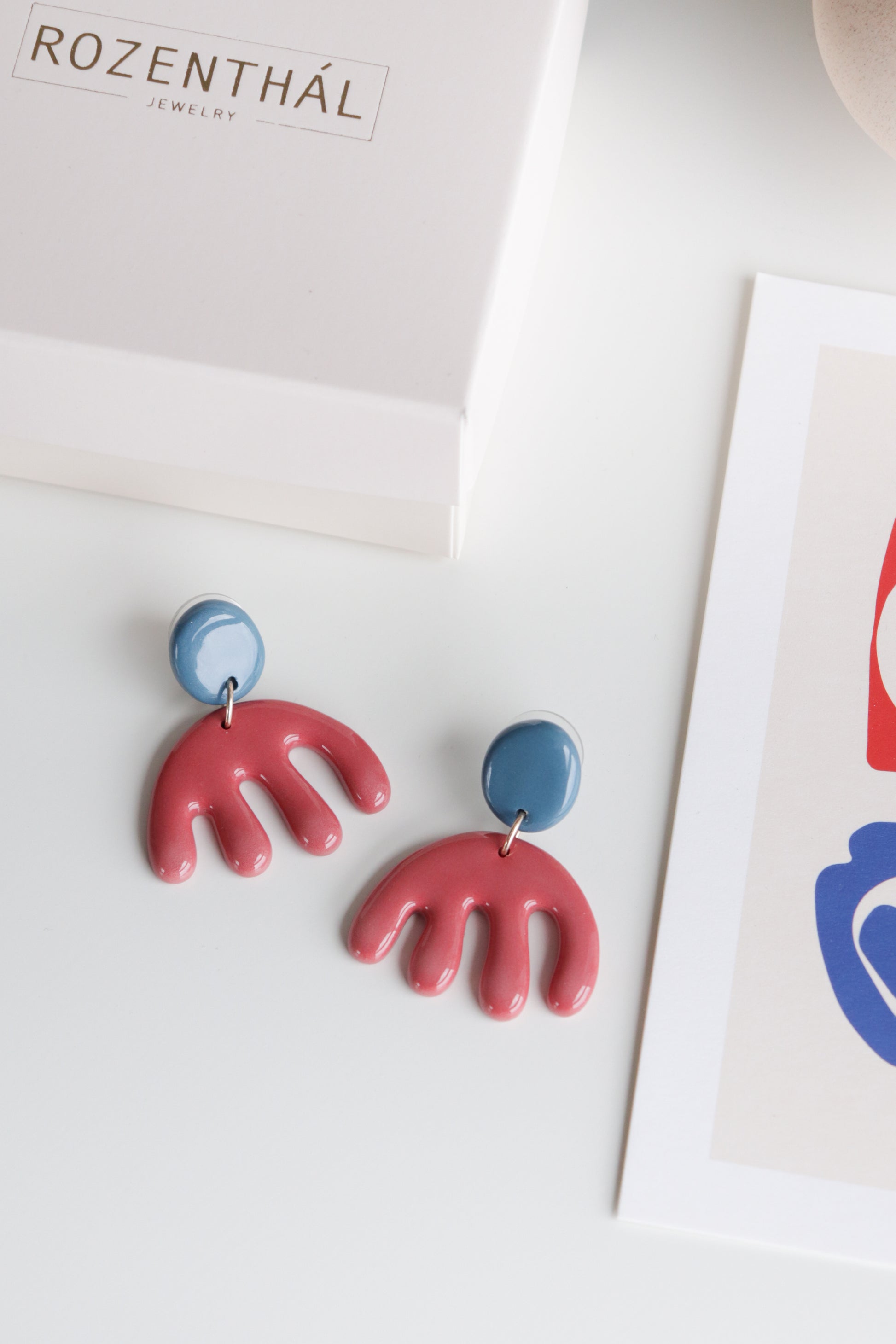 A pair of Matisse Earrings by Rozenthal Jewelry, featuring blue circular tops and red abstract hand shapes, displayed on a white surface near a branded box and an art print.