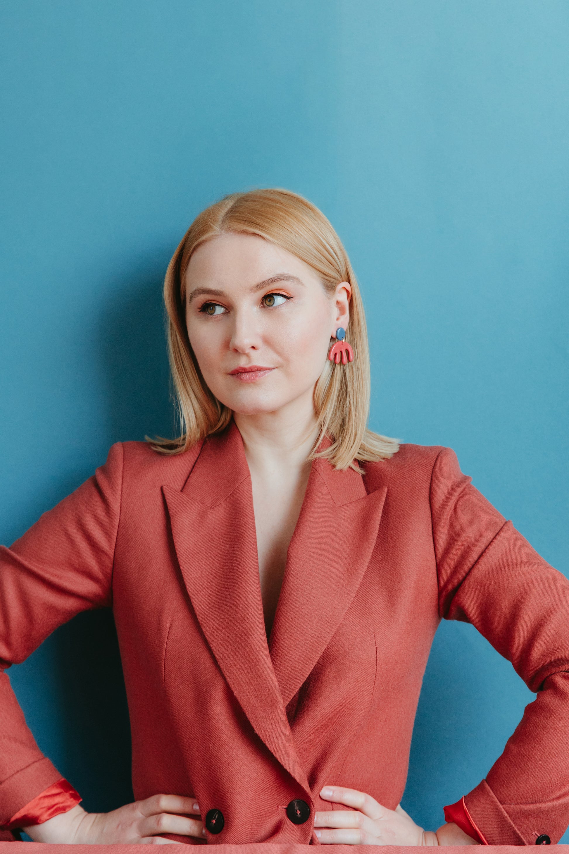 A woman with blonde hair wears Rozenthal Jewelry’s Matisse Earrings and a red blazer, standing against a blue background with her hands on her hips and a slight smile.