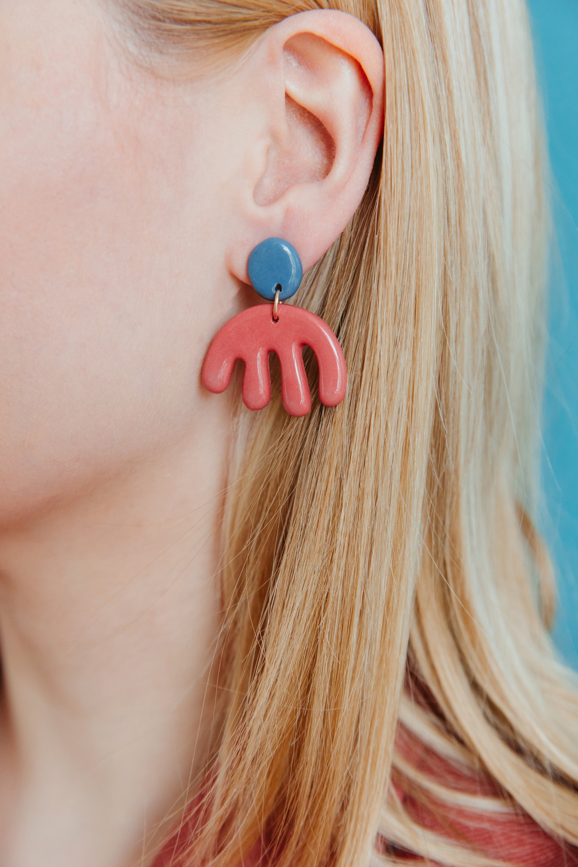 Close-up of a person with long blonde hair wearing Rozenthal Jewelry’s handmade Matisse Earrings—lightweight, waterproof, and crafted in Latvia, with a blue round stud and red arch pendant—standing out against a teal background.