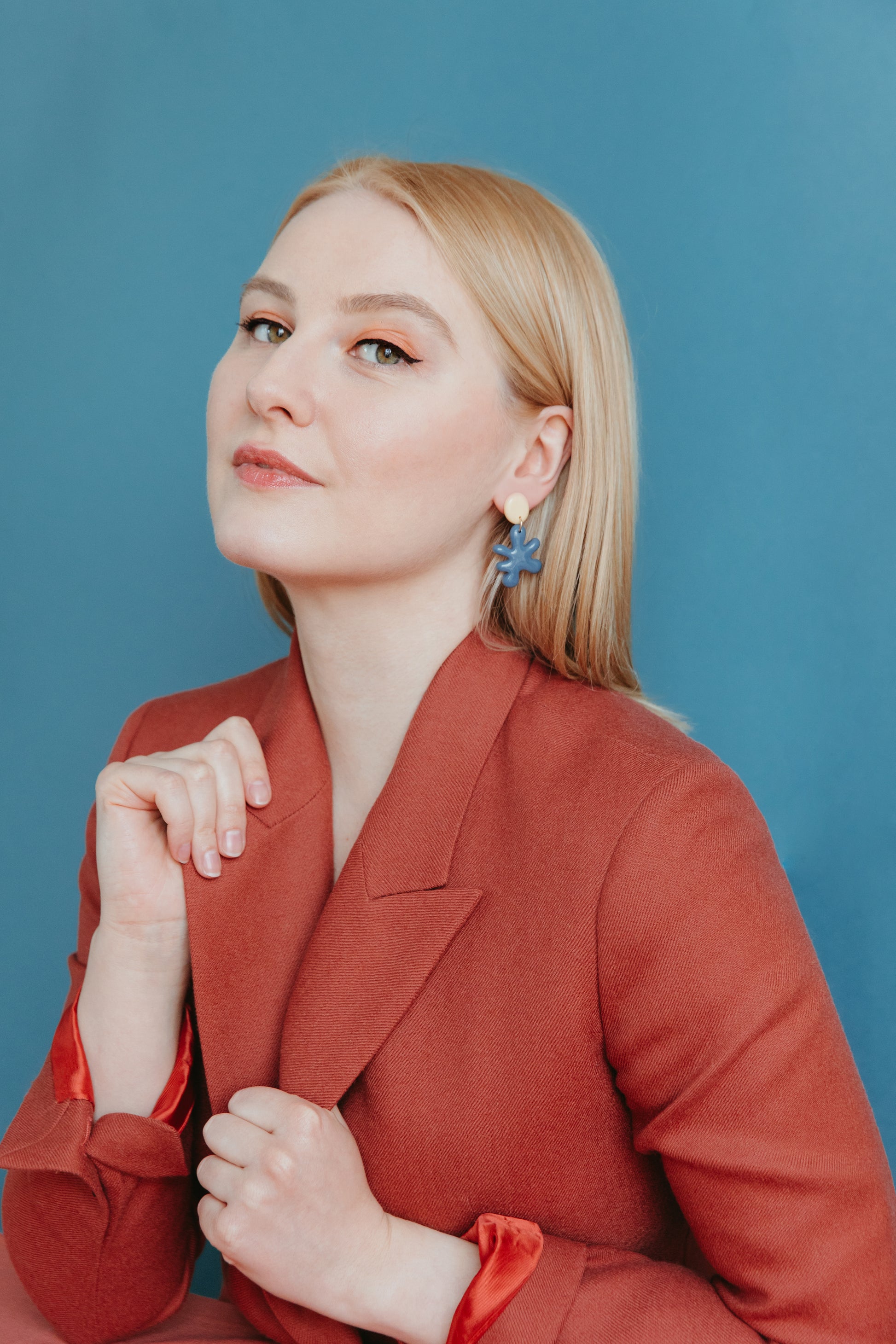 A woman with straight blonde hair confidently poses against a solid blue background, wearing Rozenthal Jewelrys handmade Matisse Earrings in blue floral porcelain and a rust-colored blazer.