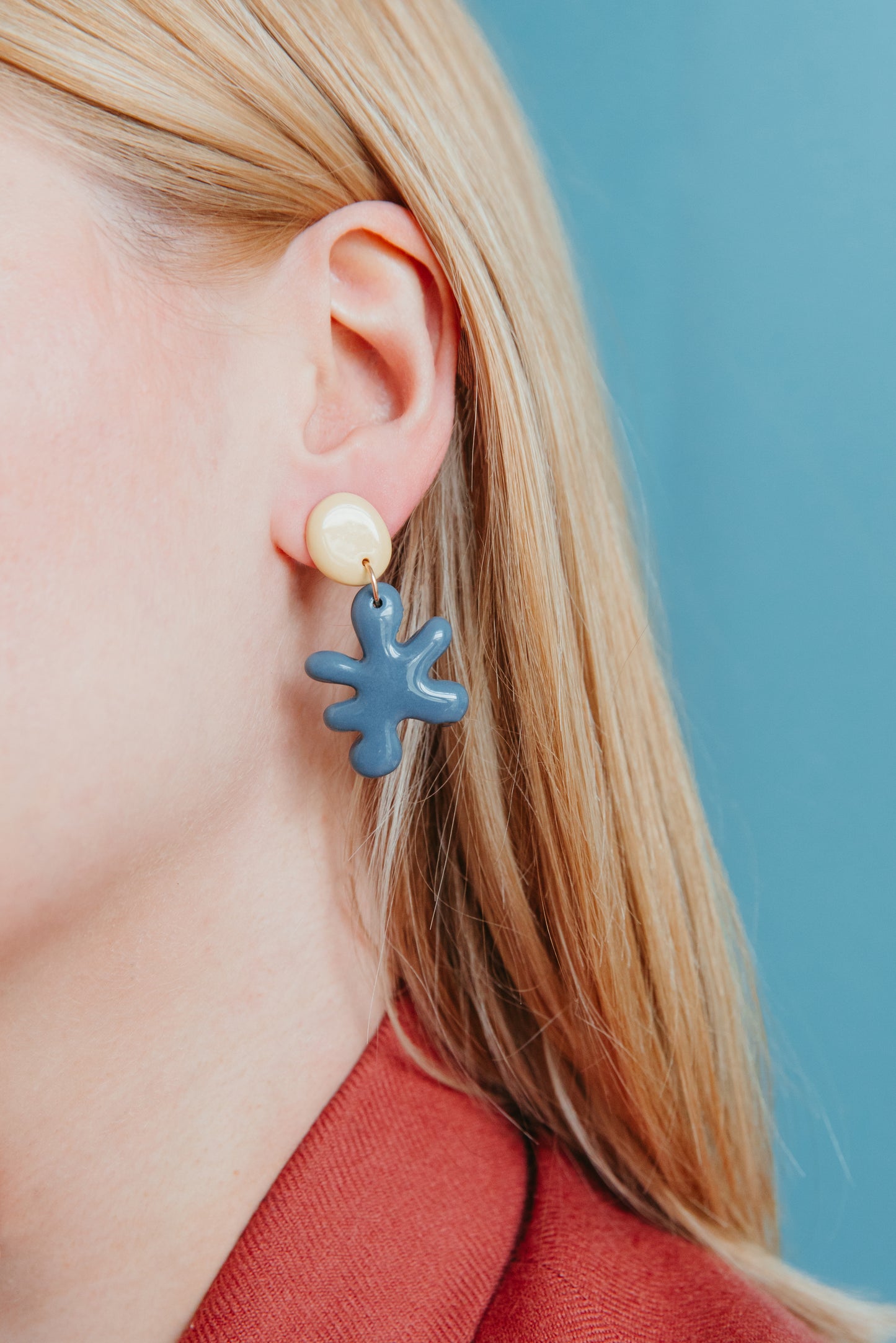 Close-up of a person with straight blonde hair in a red top, wearing Rozenthal Jewelrys Matisse Earrings—porcelain statement earrings with a round cream stud and dangling abstract blue shape—set against a blue background.