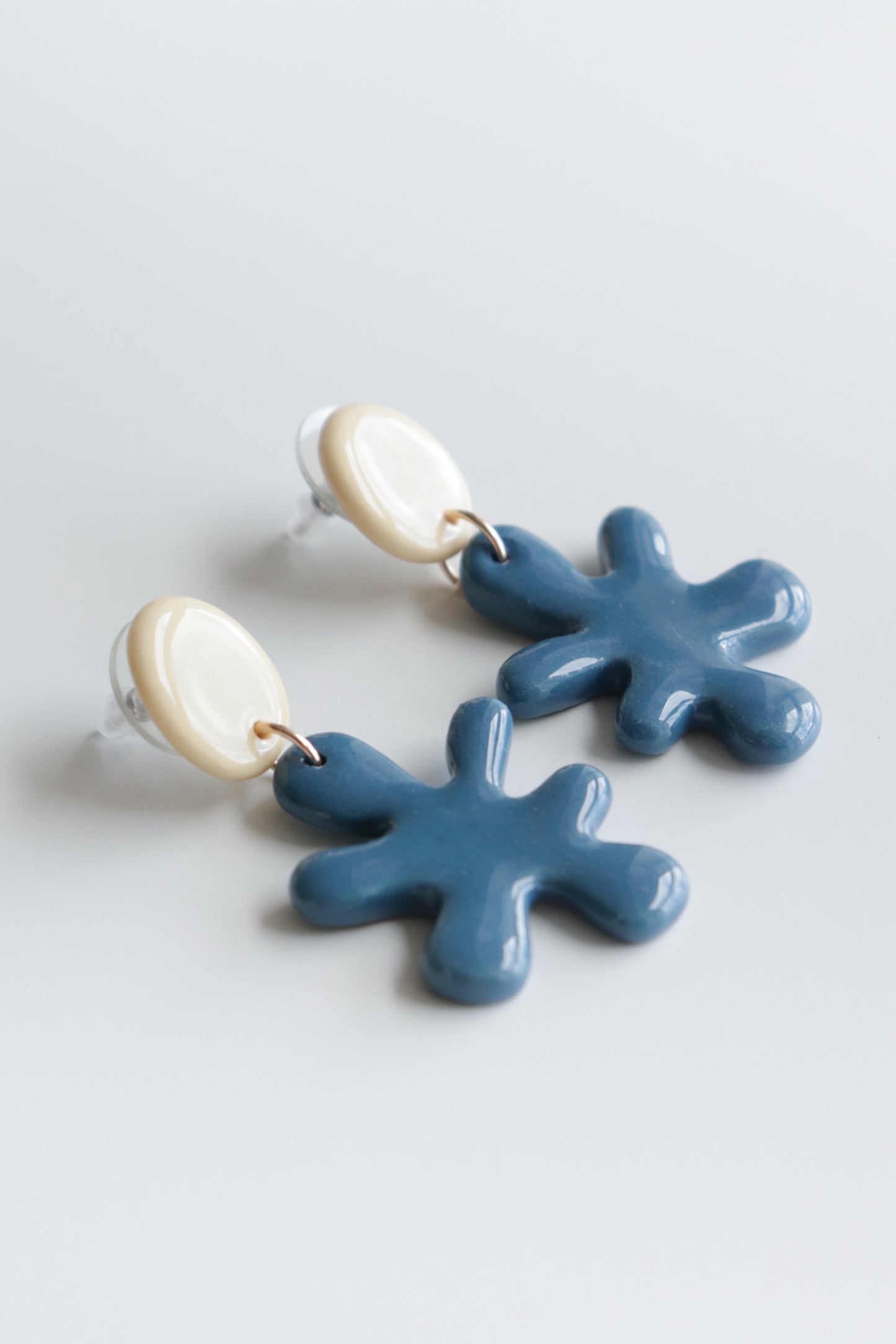 The Matisse Earrings by Rozenthal Jewelry are handmade in Latvia, with cream oval studs and dangling blue ceramic shapes resembling abstract splashes or flowers, shown on a light background.