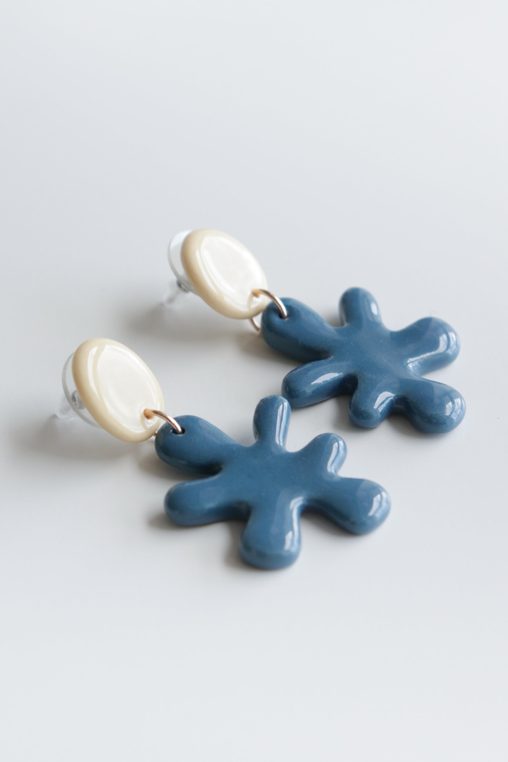 The Matisse Earrings by Rozenthal Jewelry are handmade in Latvia, with cream oval studs and dangling blue ceramic shapes resembling abstract splashes or flowers, shown on a light background.