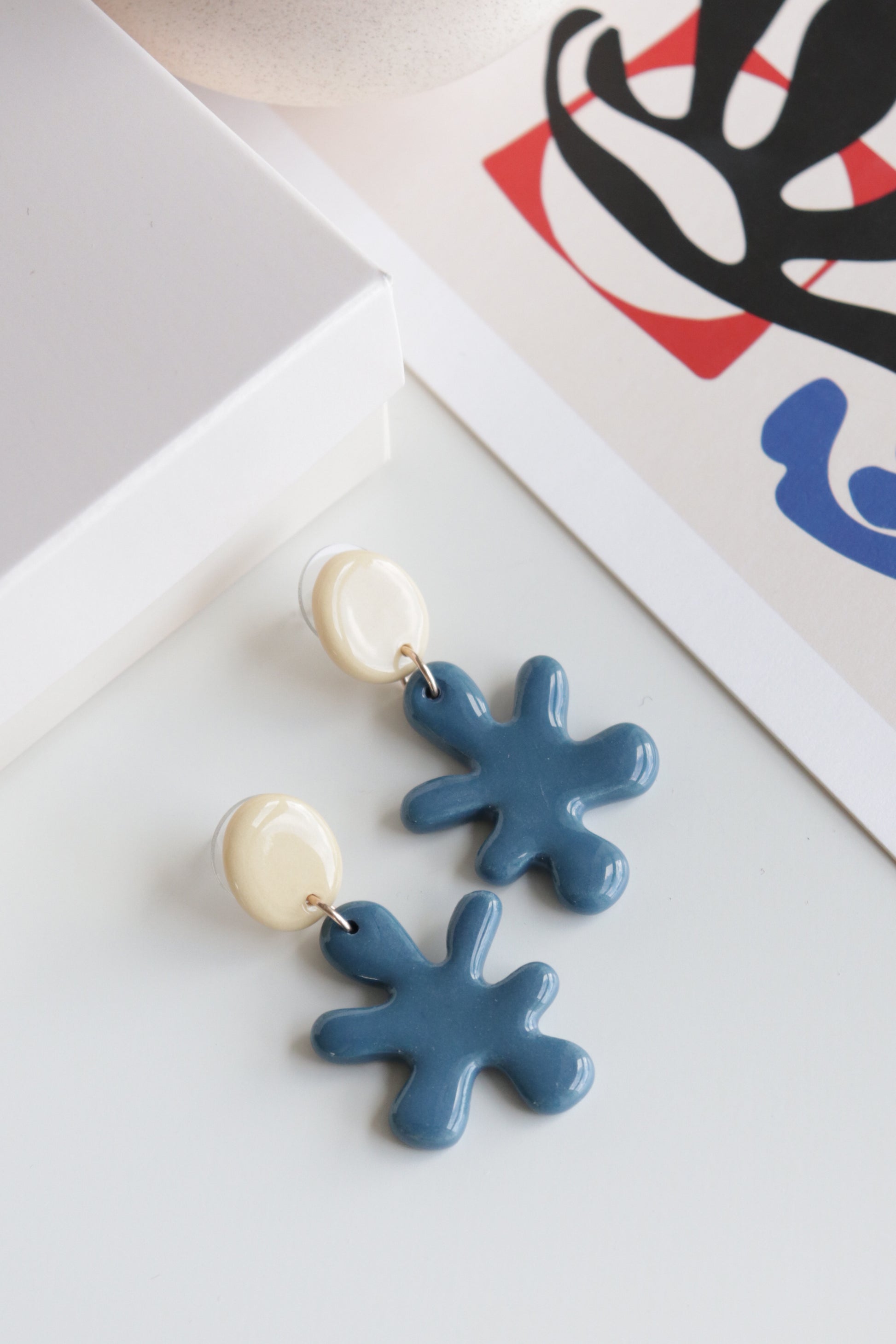 The Matisse Earrings by Rozenthal Jewelry are handmade in Latvia from porcelain, featuring cream tops and abstract blue pendants. Shown on a white surface beside a white box and a modern art print with bold colors.