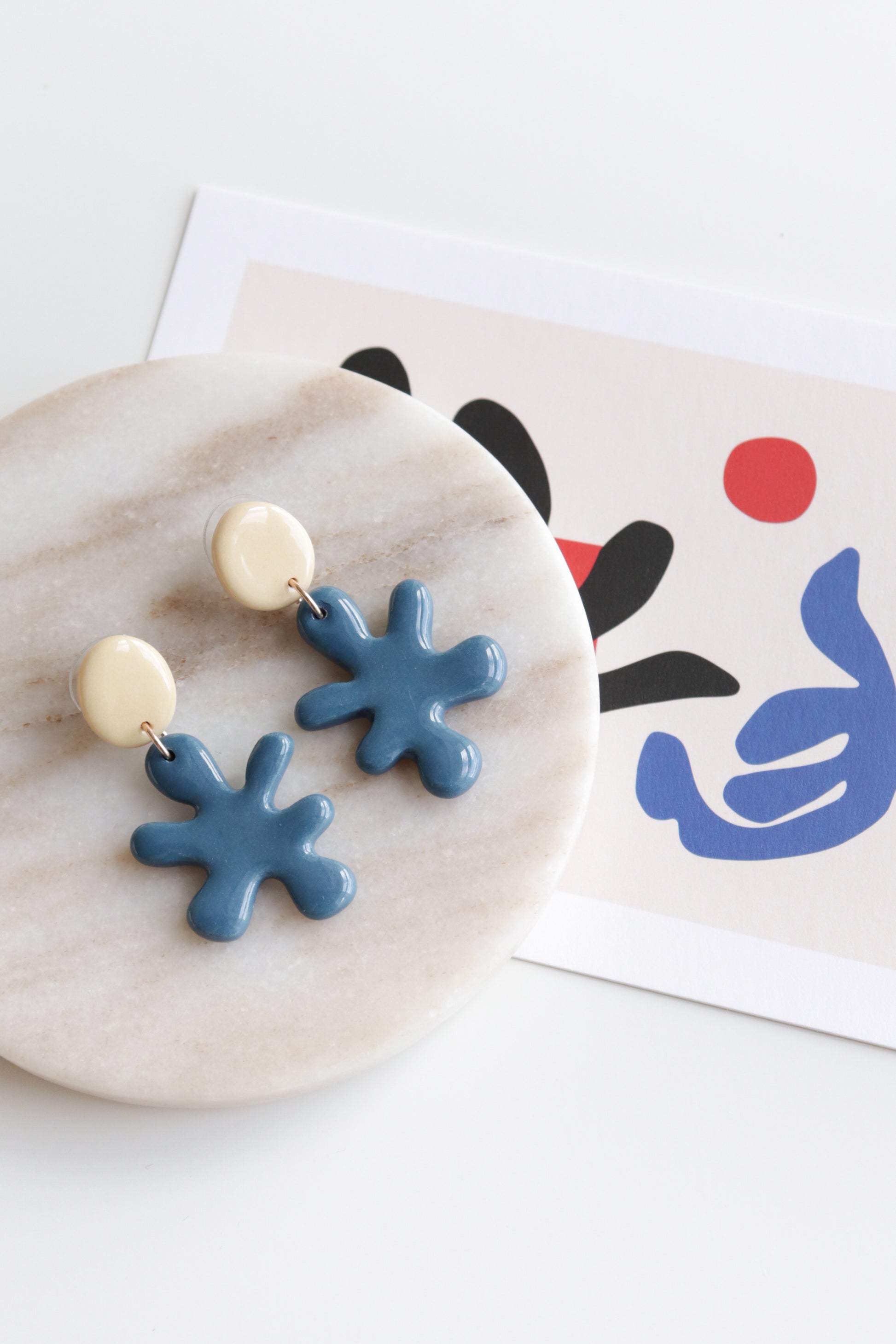 The Rozenthal Jewelry Matisse Earrings, blue abstract porcelain with cream studs, rest on a marble plate next to modern art with bold black, red, and blue shapes on white paper.