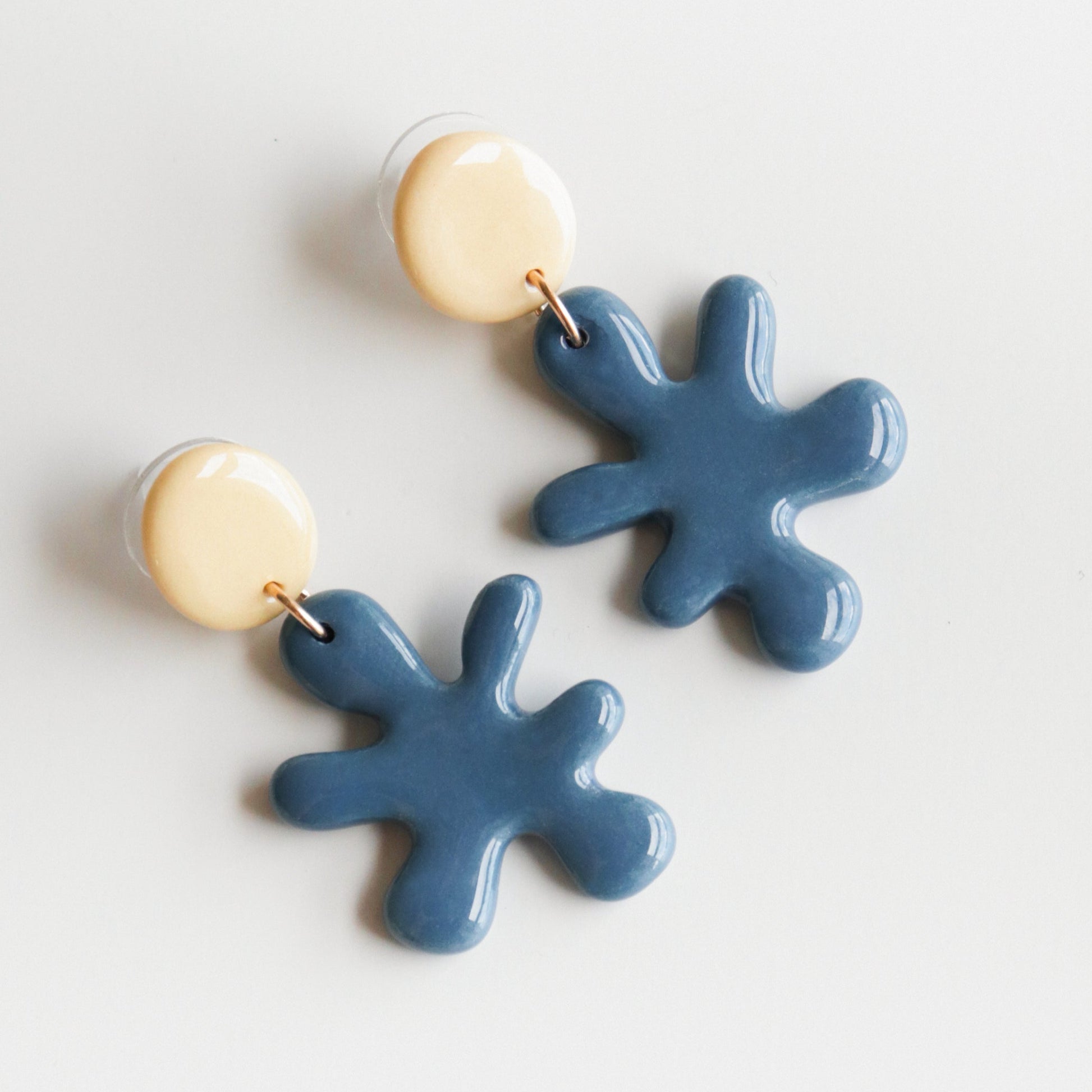 The Matisse Earrings by Rozenthal Jewelry feature round, cream-colored studs with dangling blue, splatter-shaped charms. Handmade in Latvia, these lightweight and waterproof earrings are shown on a white background.