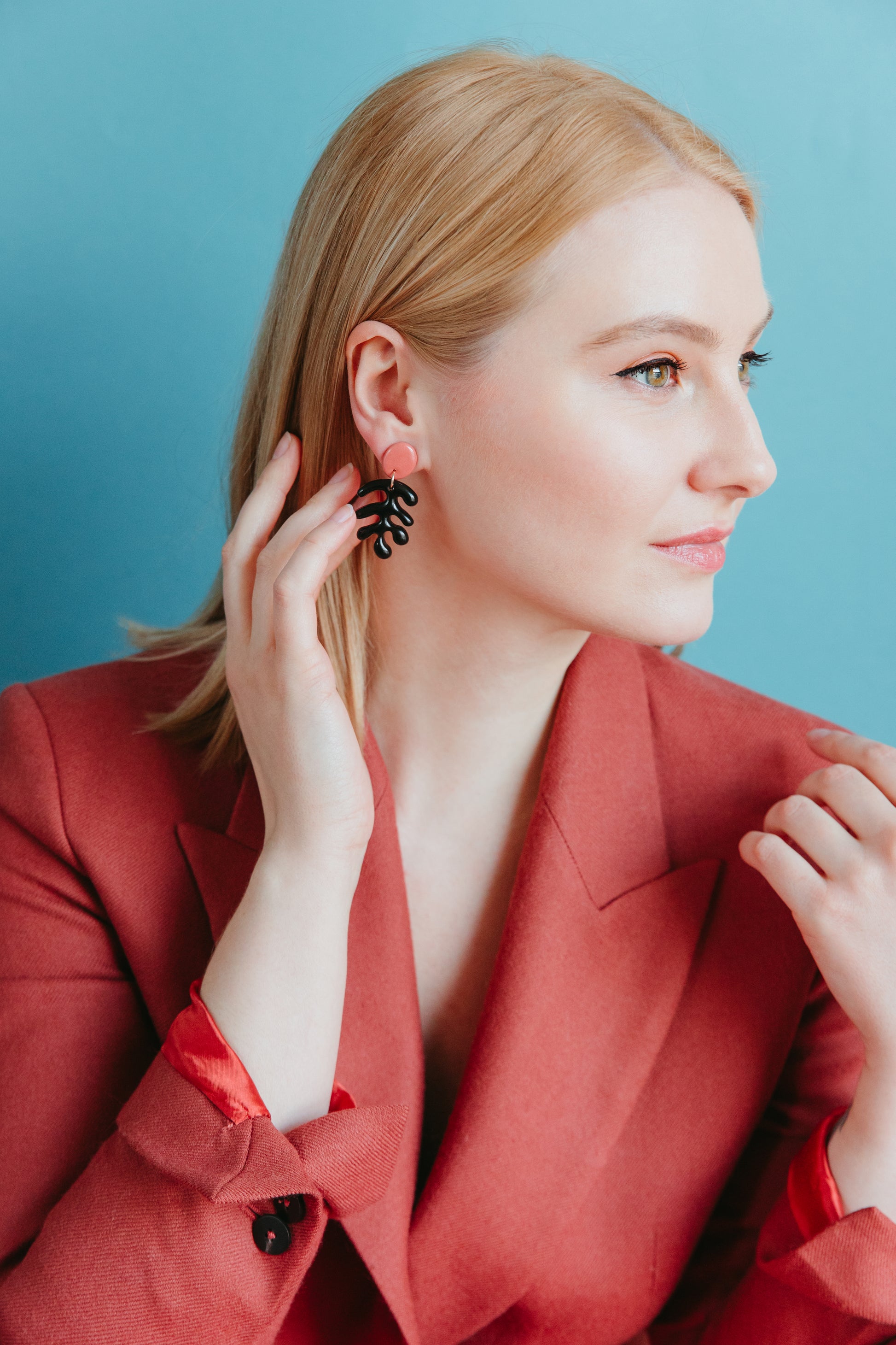 A blonde woman in a red blazer poses against a blue background, thoughtfully touching a black handmade Matisse Earring by Rozenthal Jewelry—part of a lightweight, waterproof collection for effortless style.
