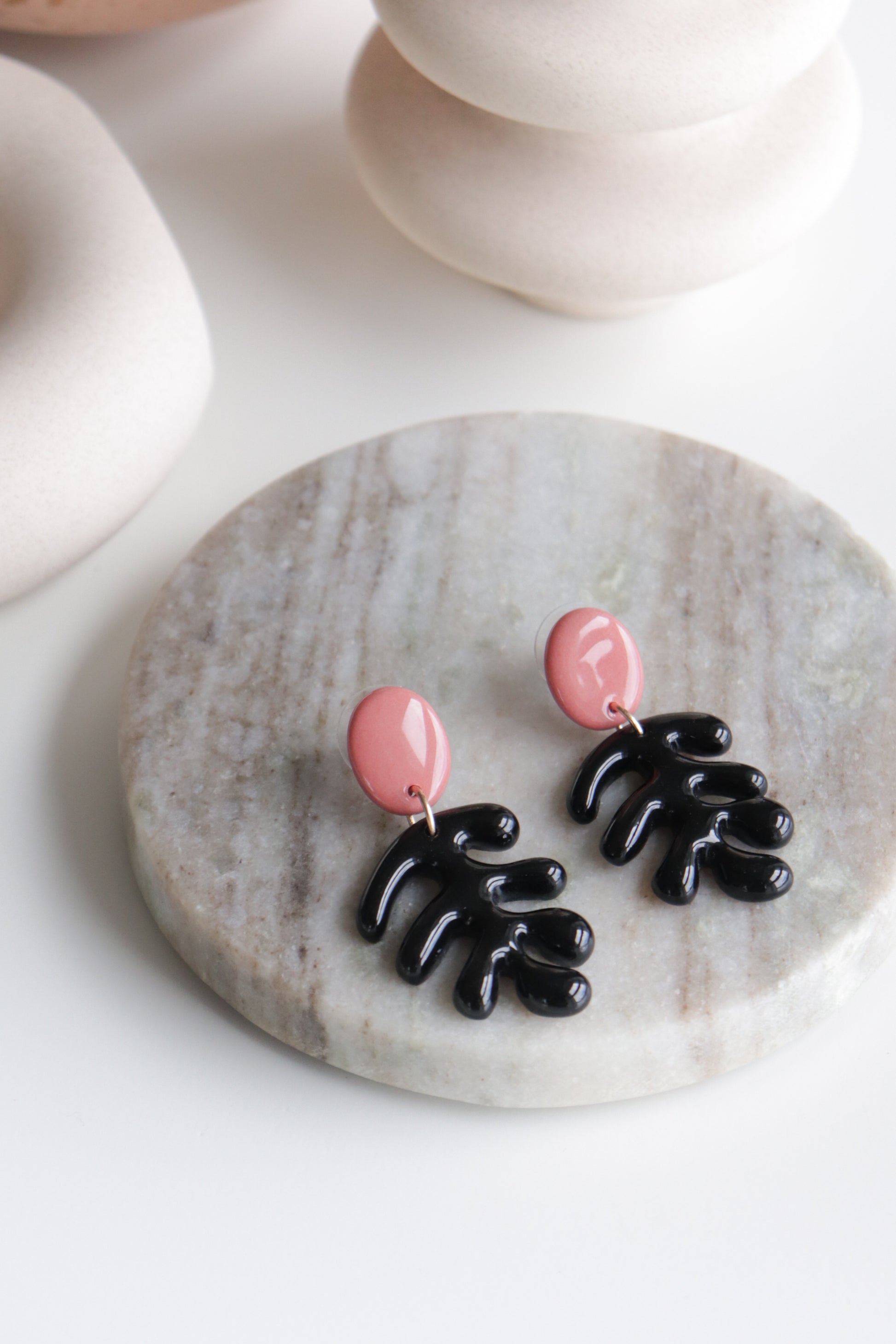 The Rozenthal Jewelry Matisse Earrings feature pink oval studs and black abstract leaf-shaped dangles. Displayed on a round marble surface, these handmade, lightweight, waterproof earrings offer effortless style.