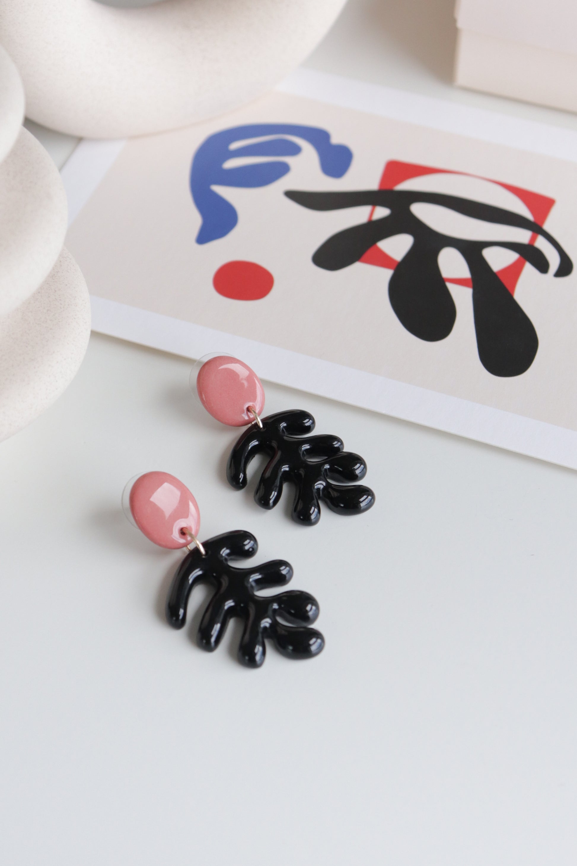 The Matisse Earrings by Rozenthal Jewelry—modern handmade earrings with pink circular studs and black abstract shapes—are displayed on a white surface, with a blue, red, and black abstract art print in the background. Curved white objects frame the scene.