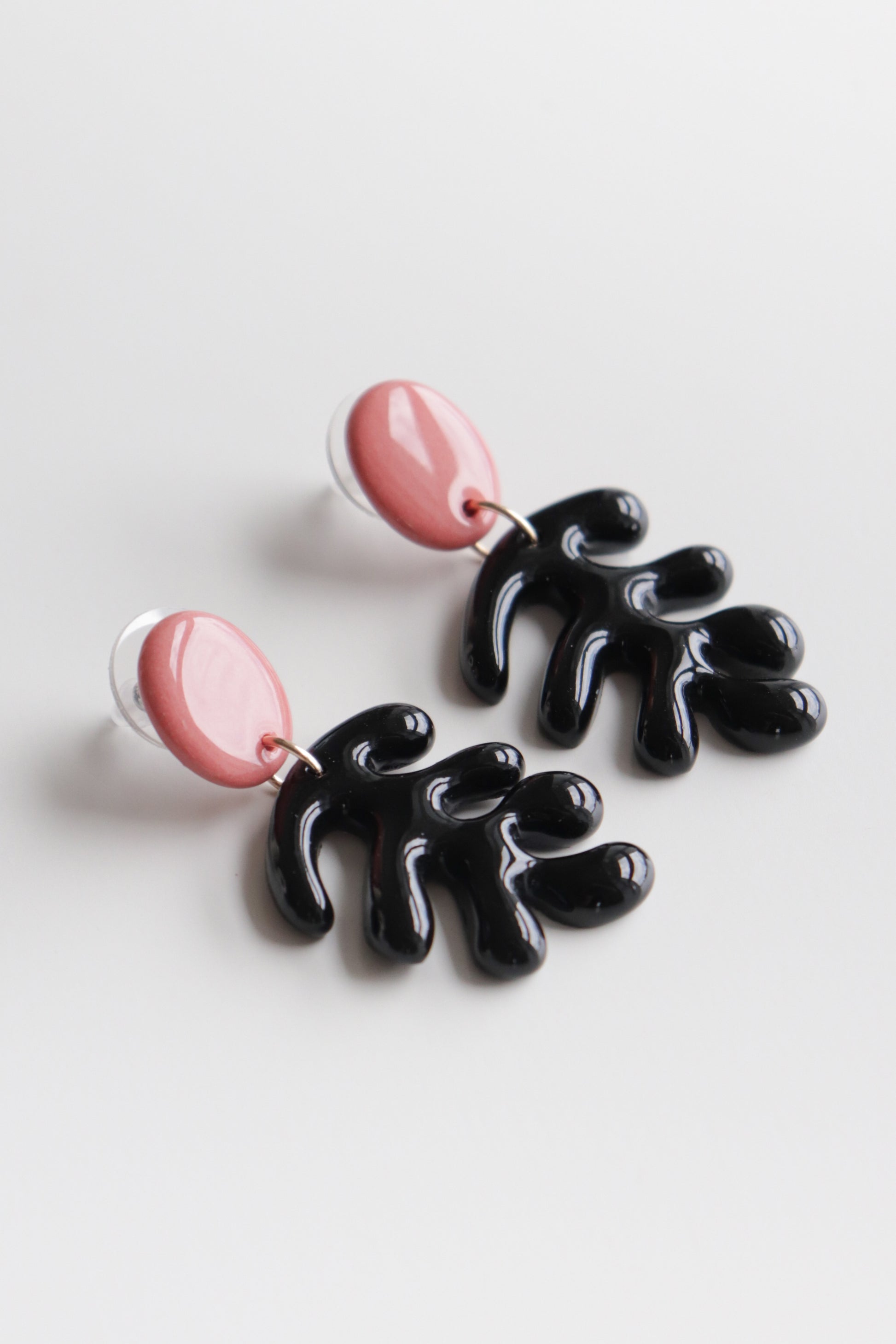 The Matisse Earrings by Rozenthal Jewelry feature glossy pink oval studs with dangling black abstract shapes, handmade in Latvia and shown on a white background.