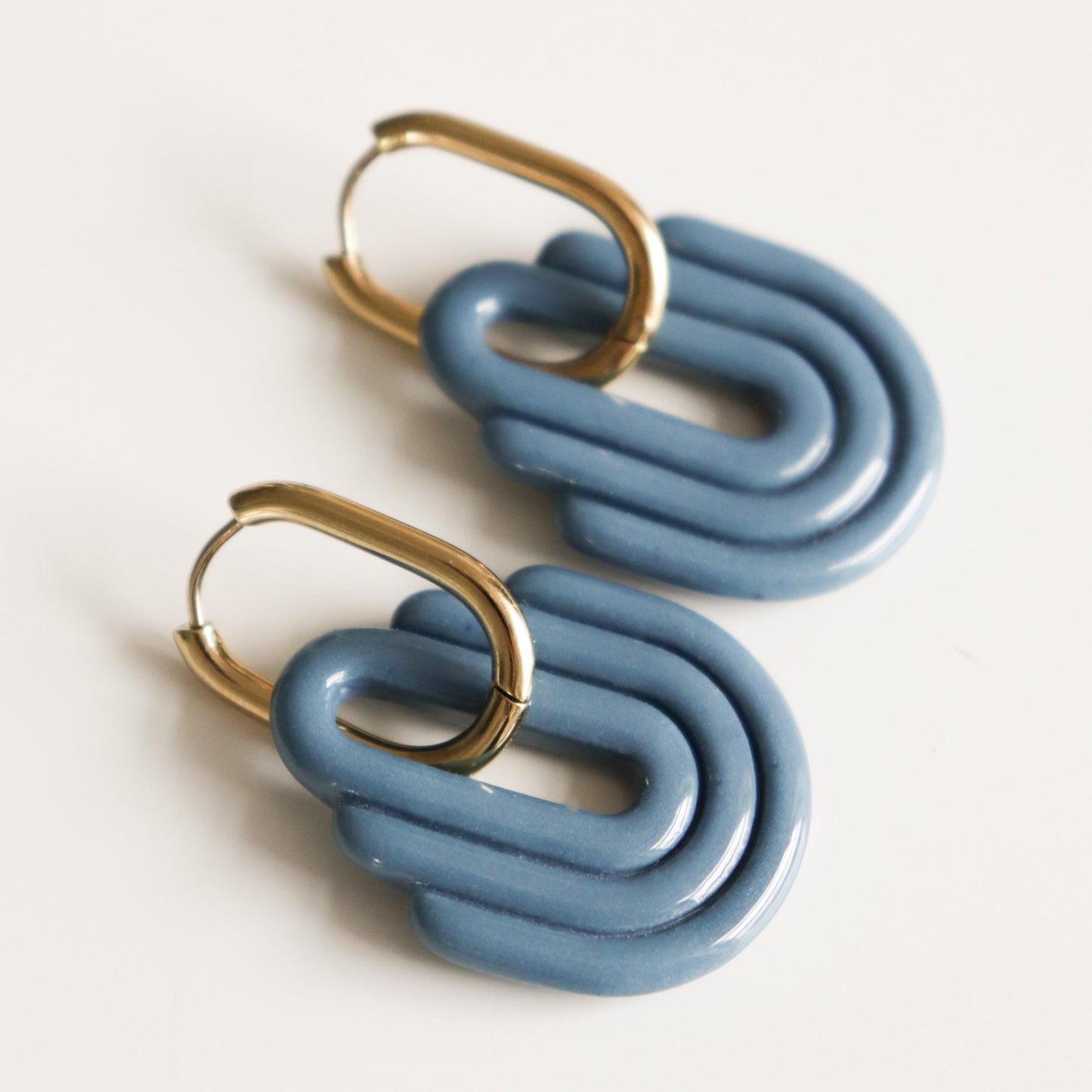 The Art Deco Earrings by Rozenthal Jewelry are hypoallergenic designer gold hoops with blue glossy interlocking geometric shapes, shown resting on a white background.