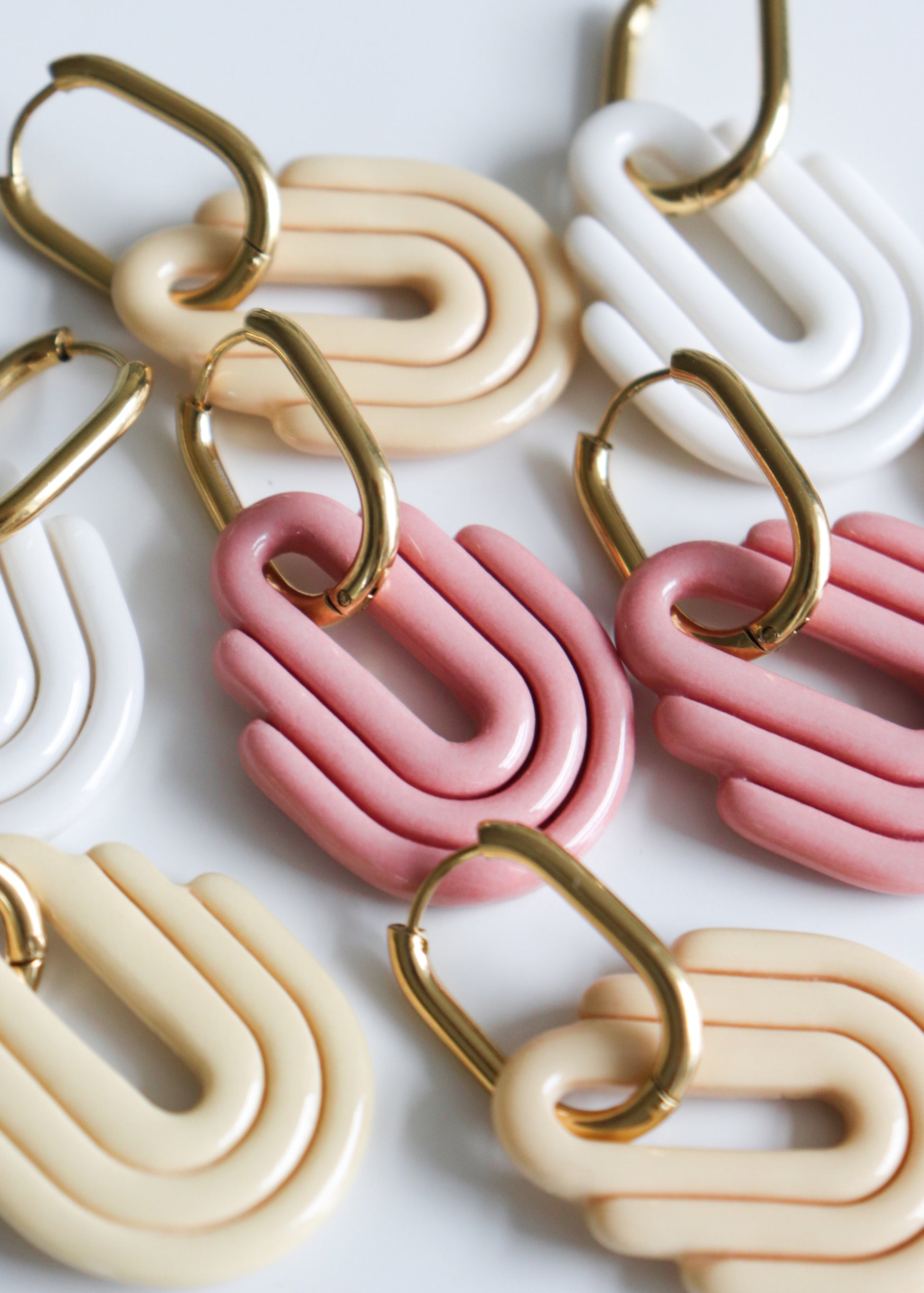 Close-up of several pairs of Art Deco Earrings by Rozenthal Jewelry with gold hooks, featuring layered U-shaped designs in cream, beige, and pink, handmade in Latvia and arranged on a white surface.