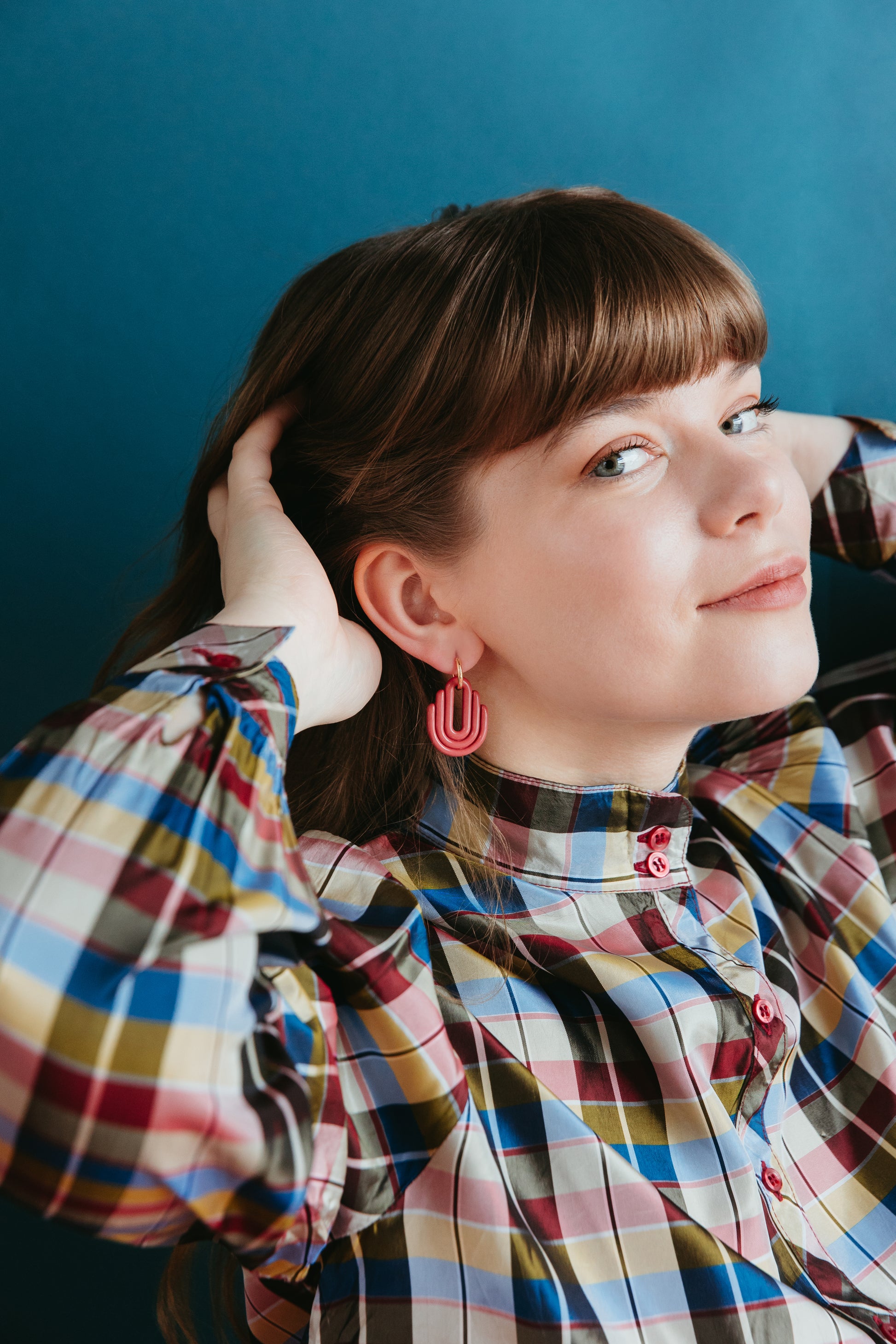 A woman with brown hair and bangs poses against a blue background, wearing a colorful plaid shirt and Rozenthal Jewelrys Art Deco Earrings, her hands raised near her head and a slight smile on her face.