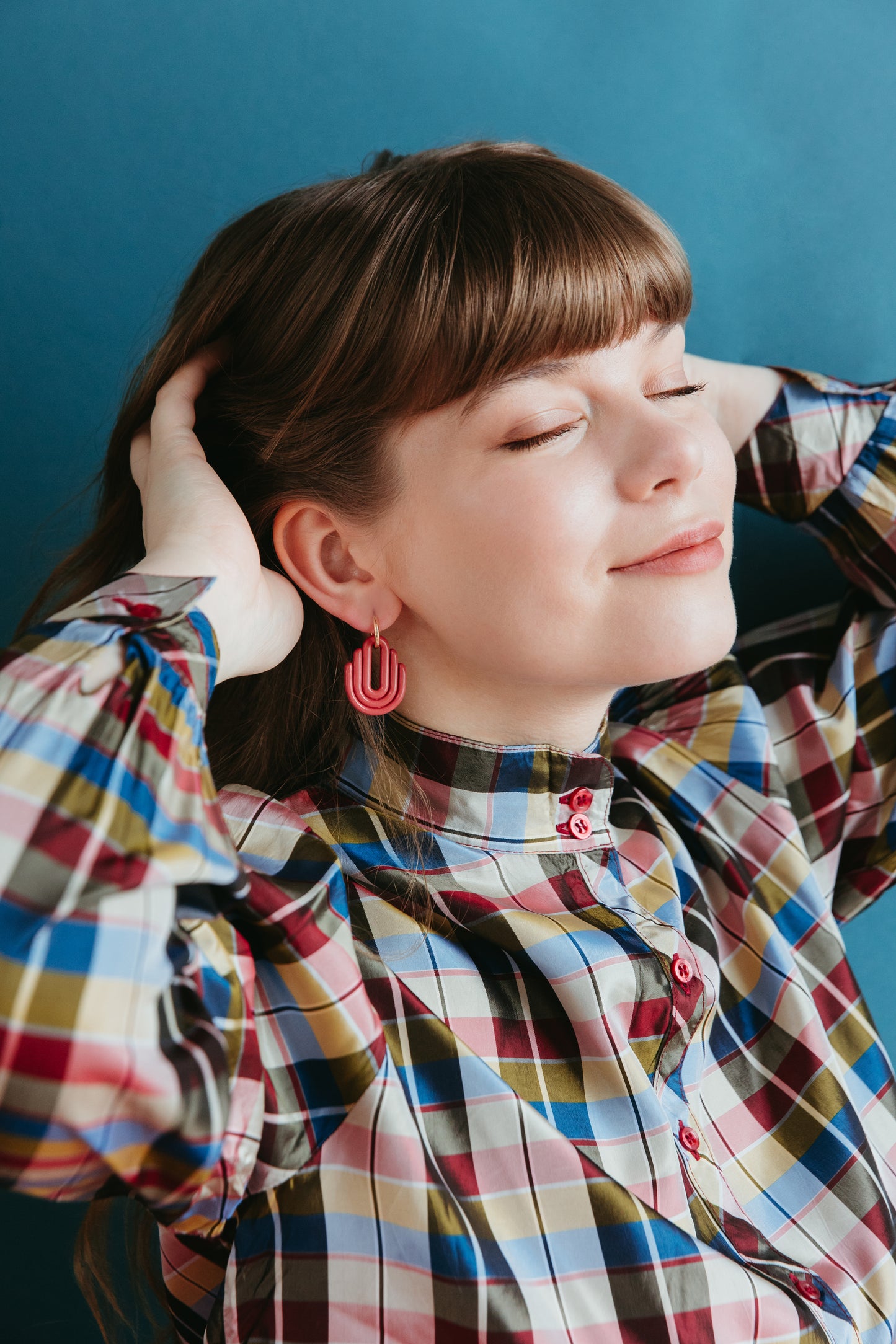 A woman with light brown hair and bangs smiles with her eyes closed, hands behind her head. She wears a colorful plaid shirt and showcases Rozenthal Jewelrys pink hypoallergenic Art Deco Earrings against a blue background.