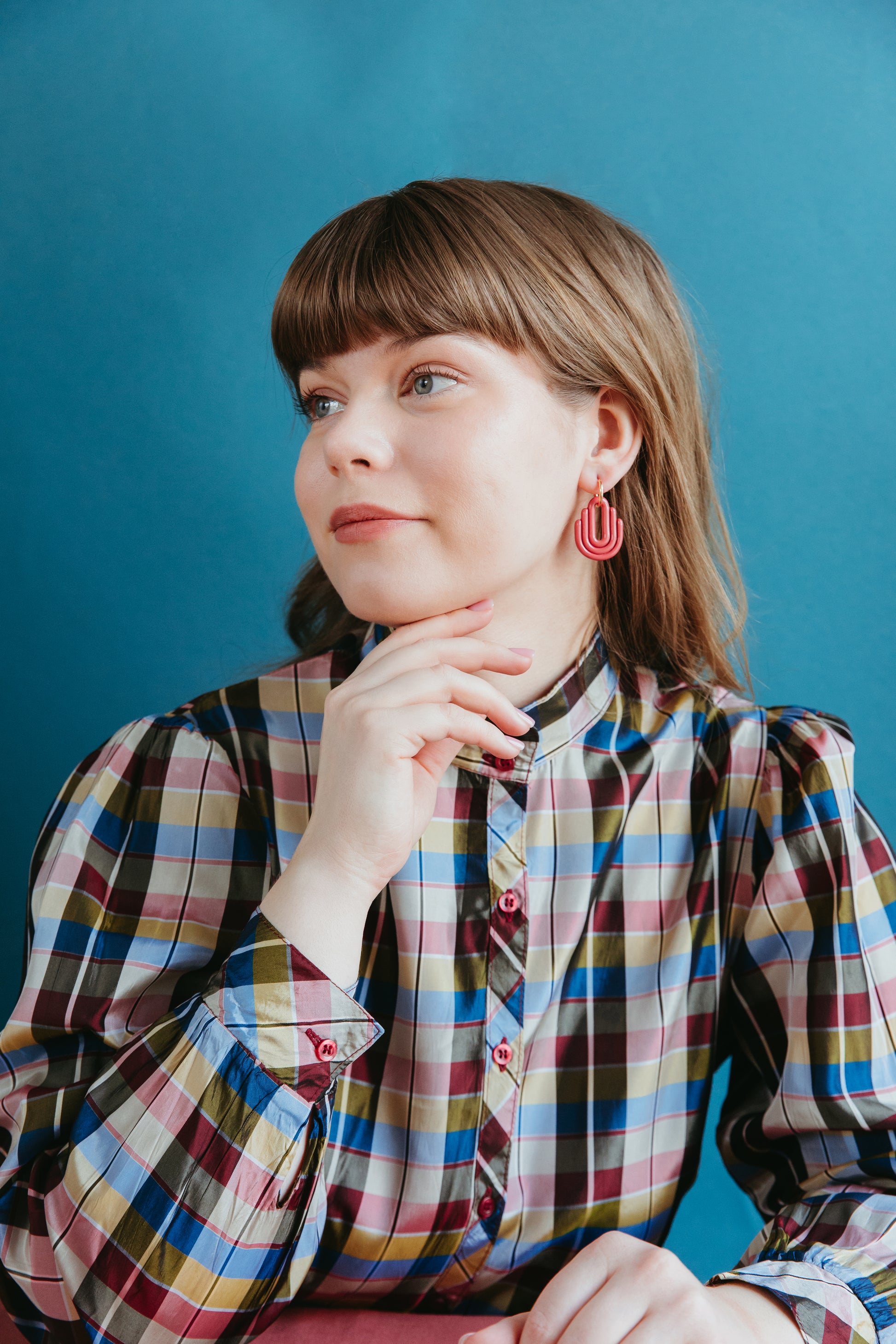 A woman with light brown hair and bangs, in a colorful plaid shirt, models Rozenthal Jewelrys Art Deco Earrings while sitting with her chin on her hand against a blue background.