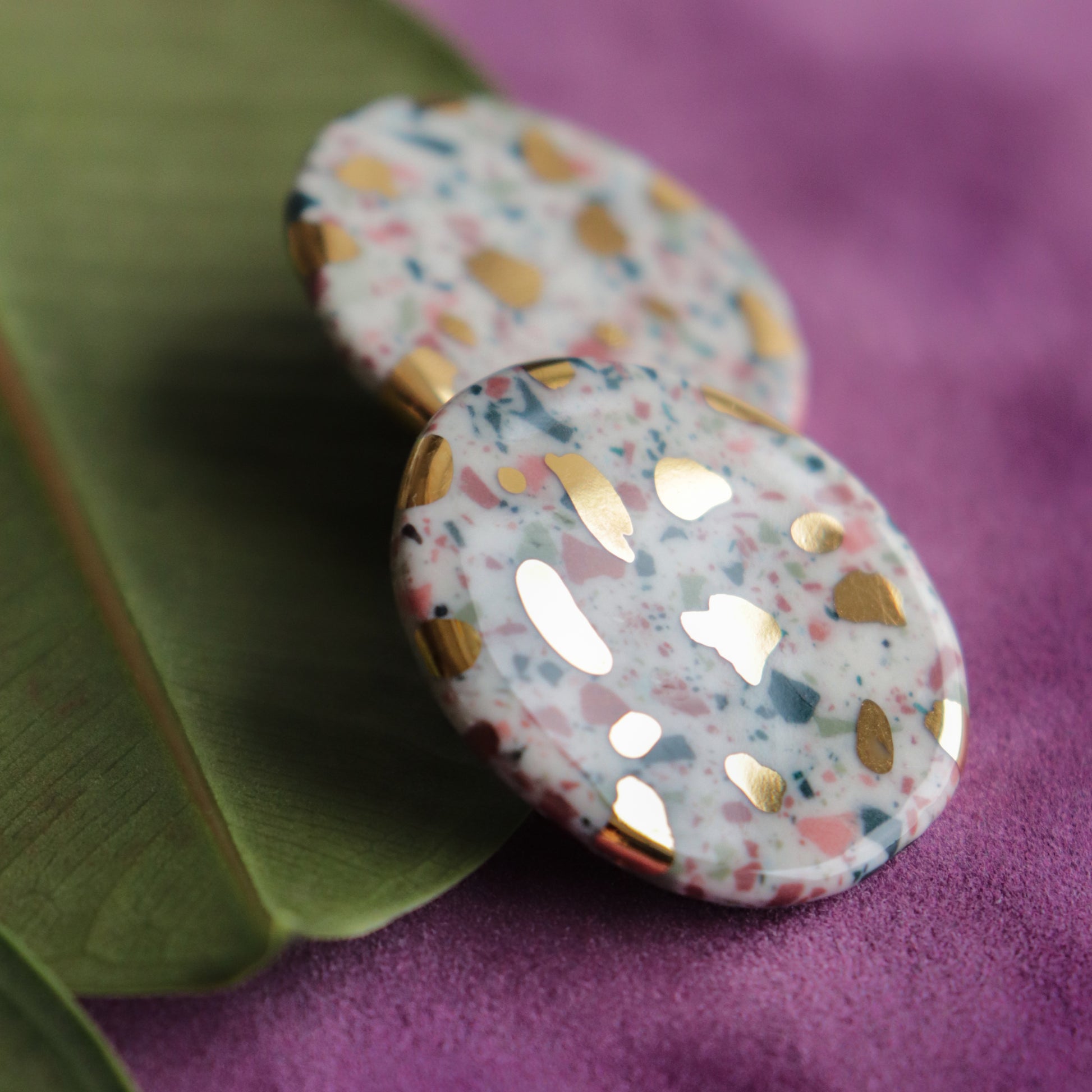 Close-up of Rozenthal Jewelrys Astilla Recycled Studs / M—round, handmade earrings with a terrazzo pattern in pink, blue, and white plus gold accents—displayed on a green leaf against a purple background.